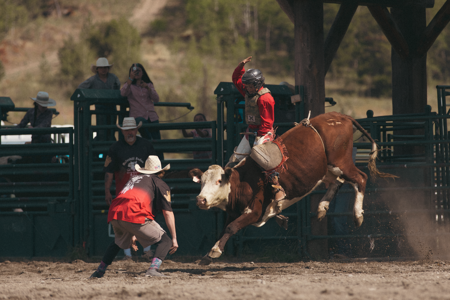 A rodeo rider in red atop a bucking bull while a clown assists at an outdoor rodeo arena.