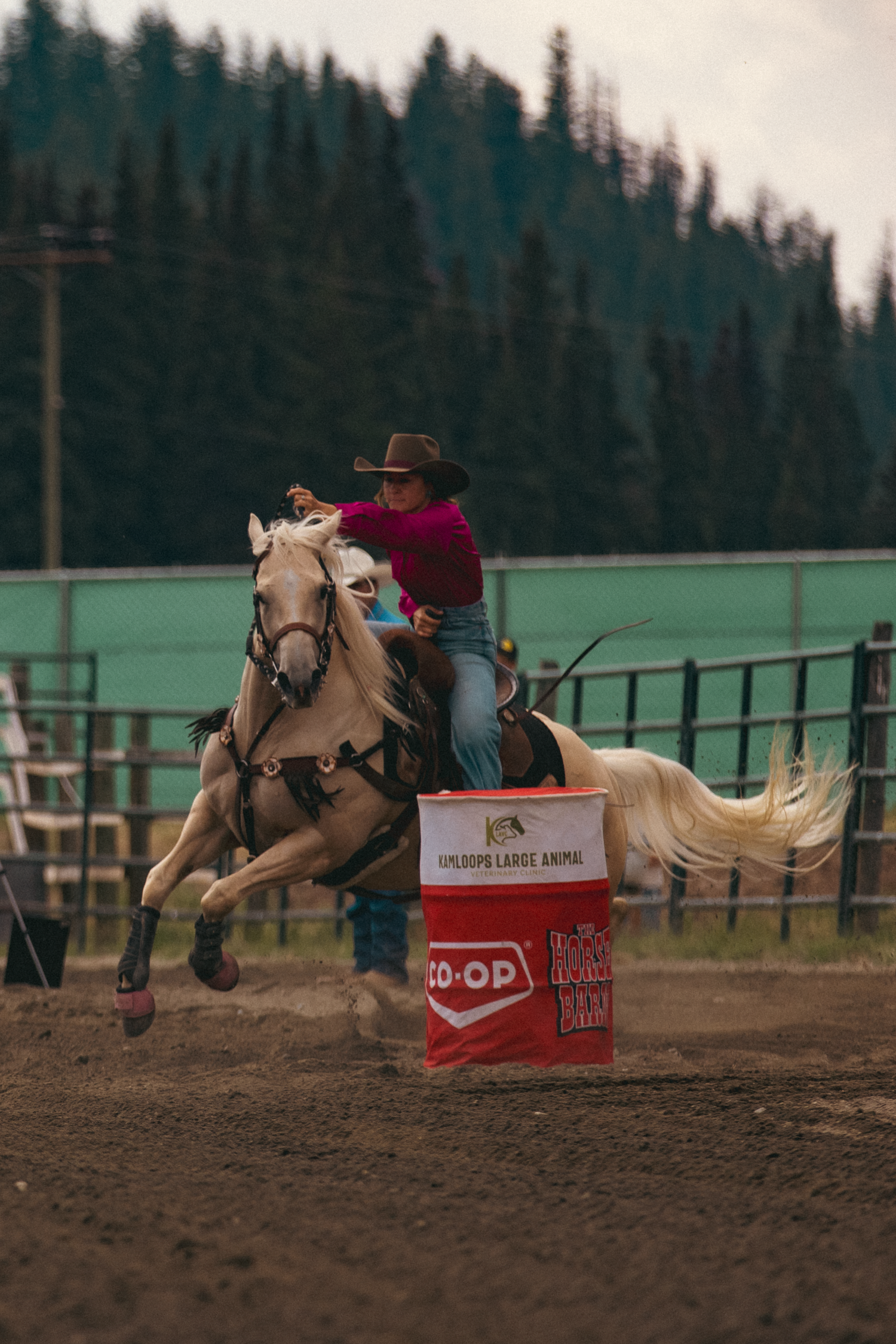 A cowgirl in a pink shirt and cowboy hat races around a barrel on a palomino horse at a rodeo competition.