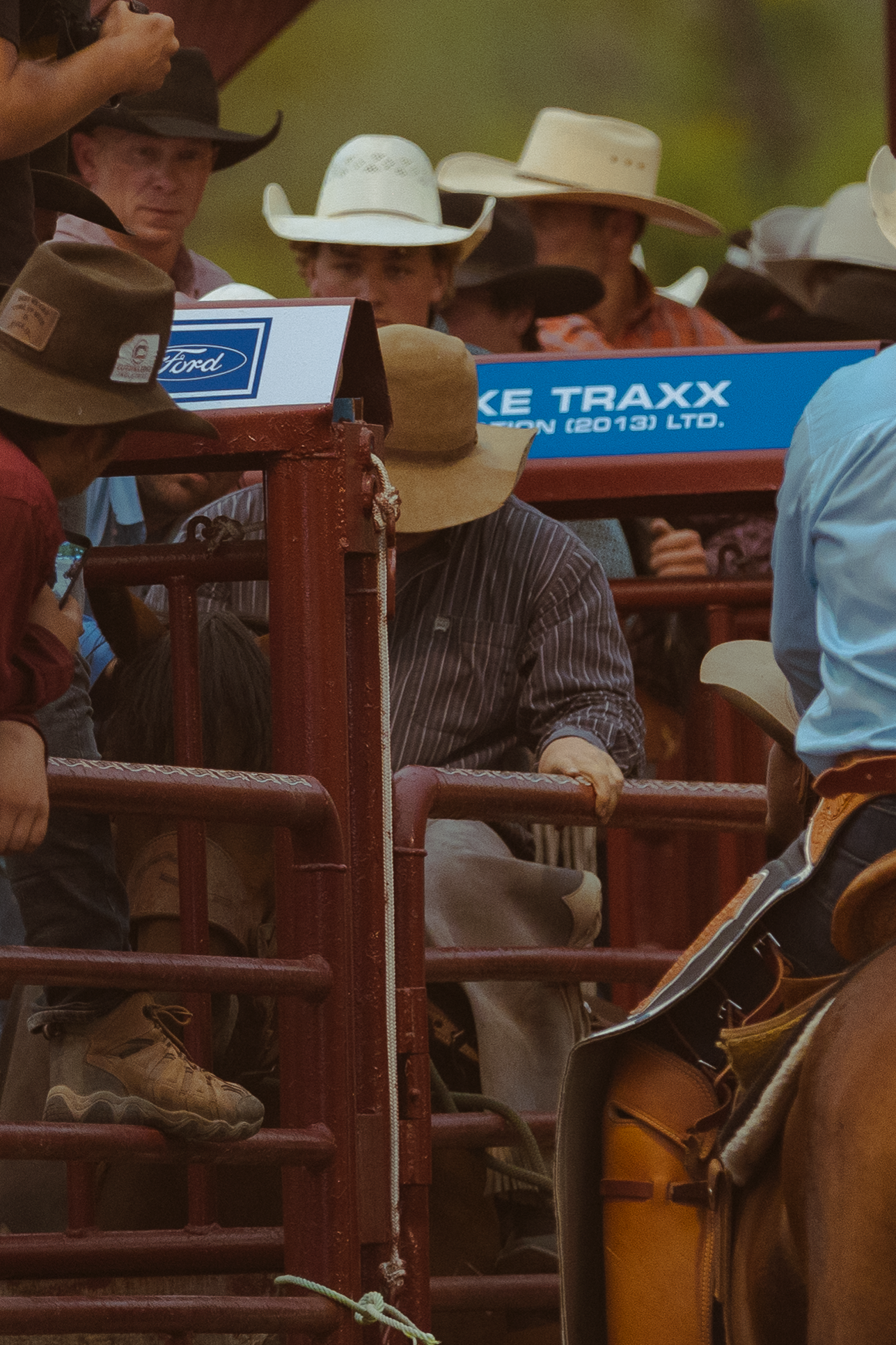Cowboys in cowboy hats crowd around a red rodeo chute with a Ford sponsor sign at a western rodeo.