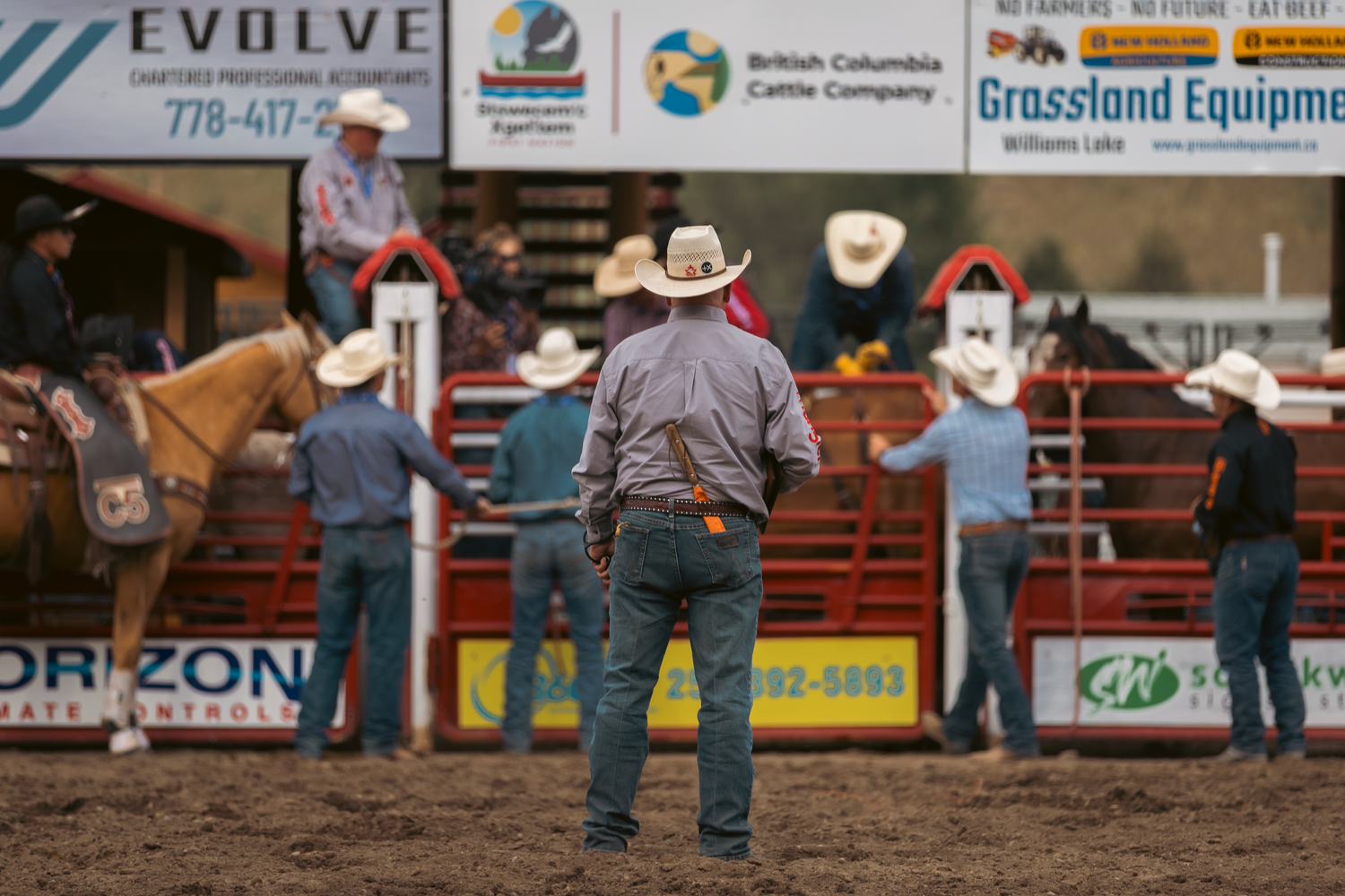 Cowboys stand in a rodeo arena with sponsor banners including Evolve and Grassland Equipment visible in the background.