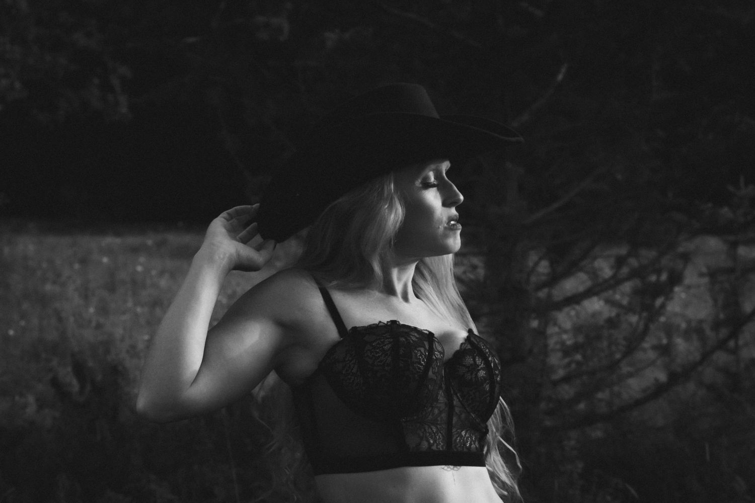 A blonde woman wearing a black wide-brim hat and lace bralette poses in a dramatic black and white portrait.