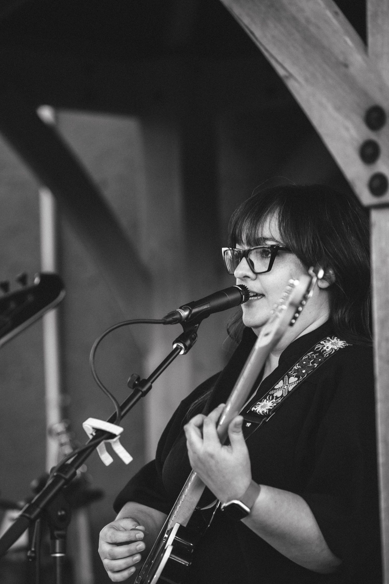 Woman with glasses singing into microphone while playing electric guitar on stage in black and white.
