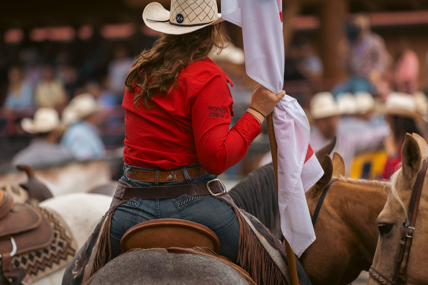 Female rodeo rider in red shirt and cowboy hat holds a flag while seated on horseback.