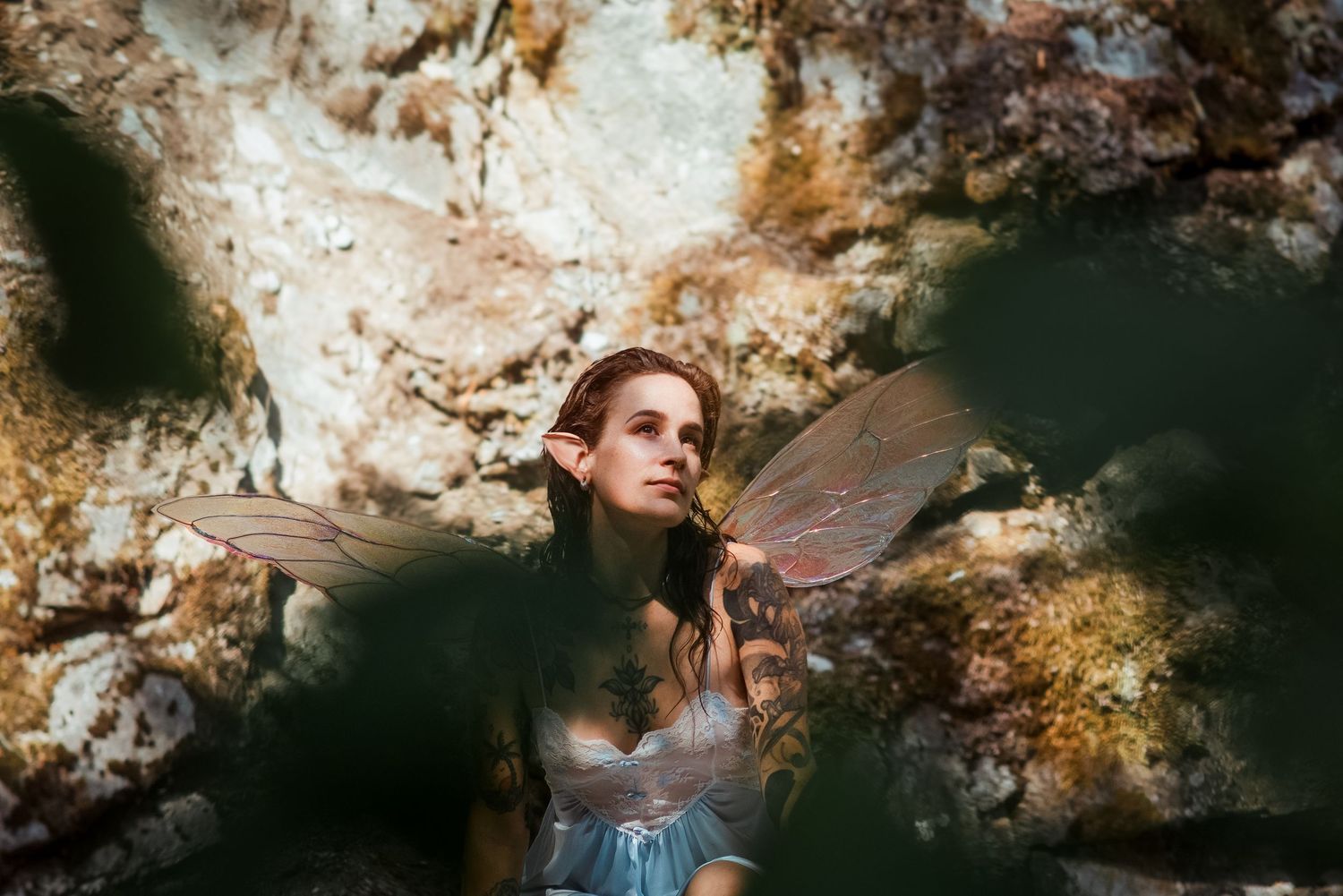 A young woman in a fairy costume with translucent wings poses among rocks in an ethereal fantasy-themed photo shoot.