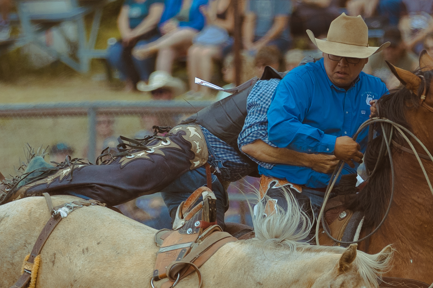 A cowboy in a blue shirt grips his rope tightly while riding a horse at a rodeo event.