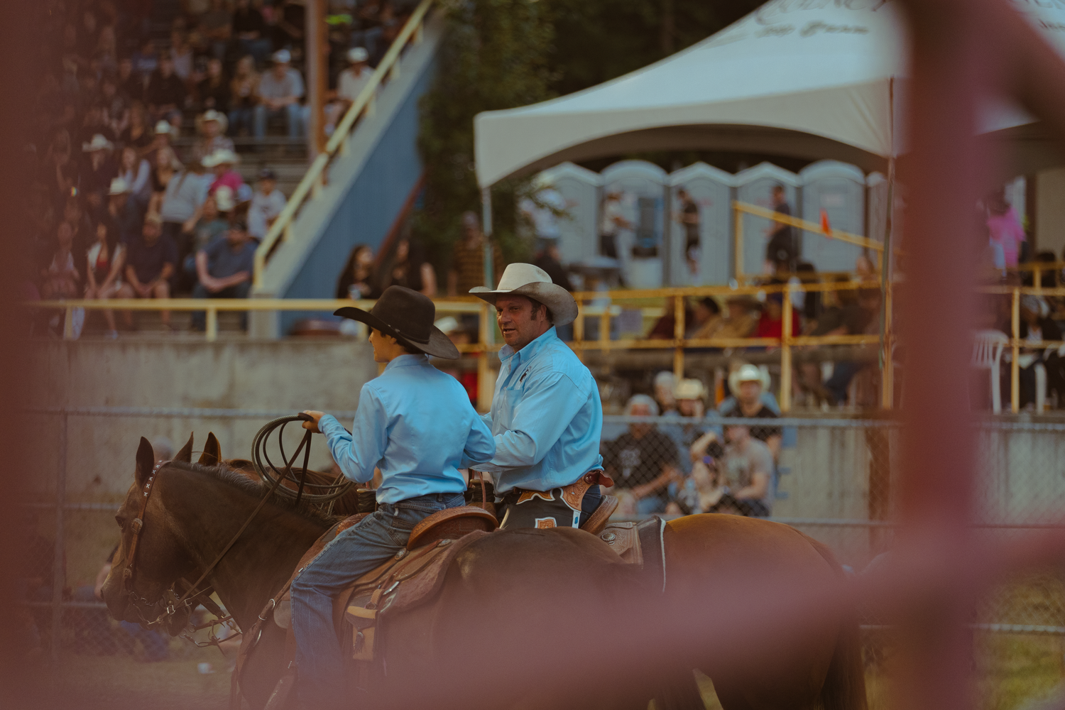 Two cowboys on horseback ride through a rodeo arena viewed through a metal fence gate.