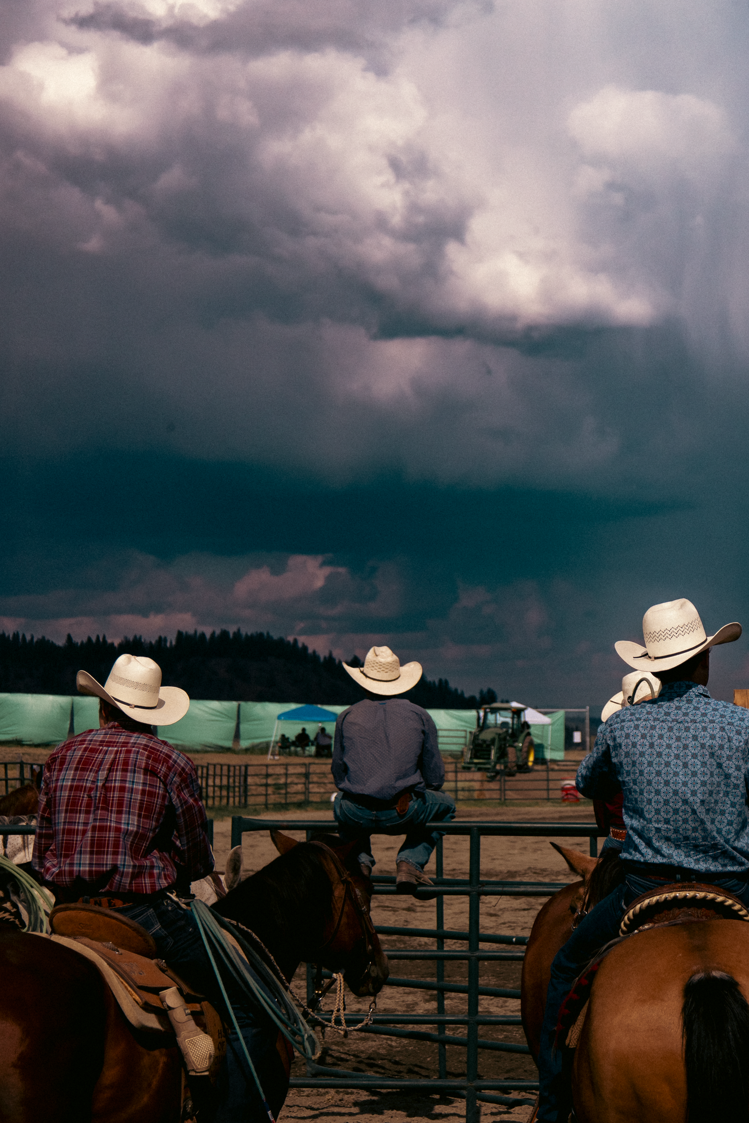 Three cowboys in white hats on horseback watch a stormy sky over a rodeo arena.