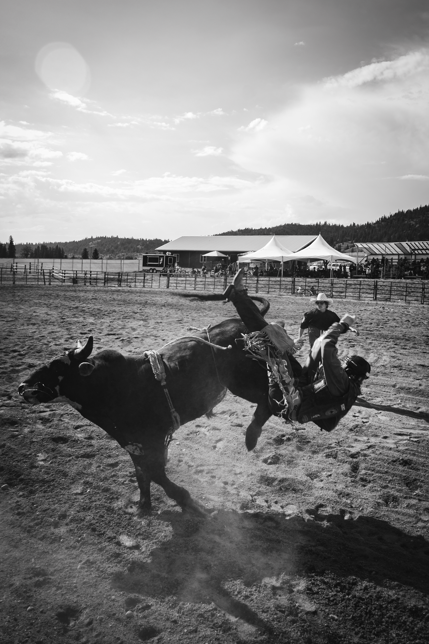 Black and white photo of a cowboy riding a bucking bull at a rodeo arena with spectator tents in the background.