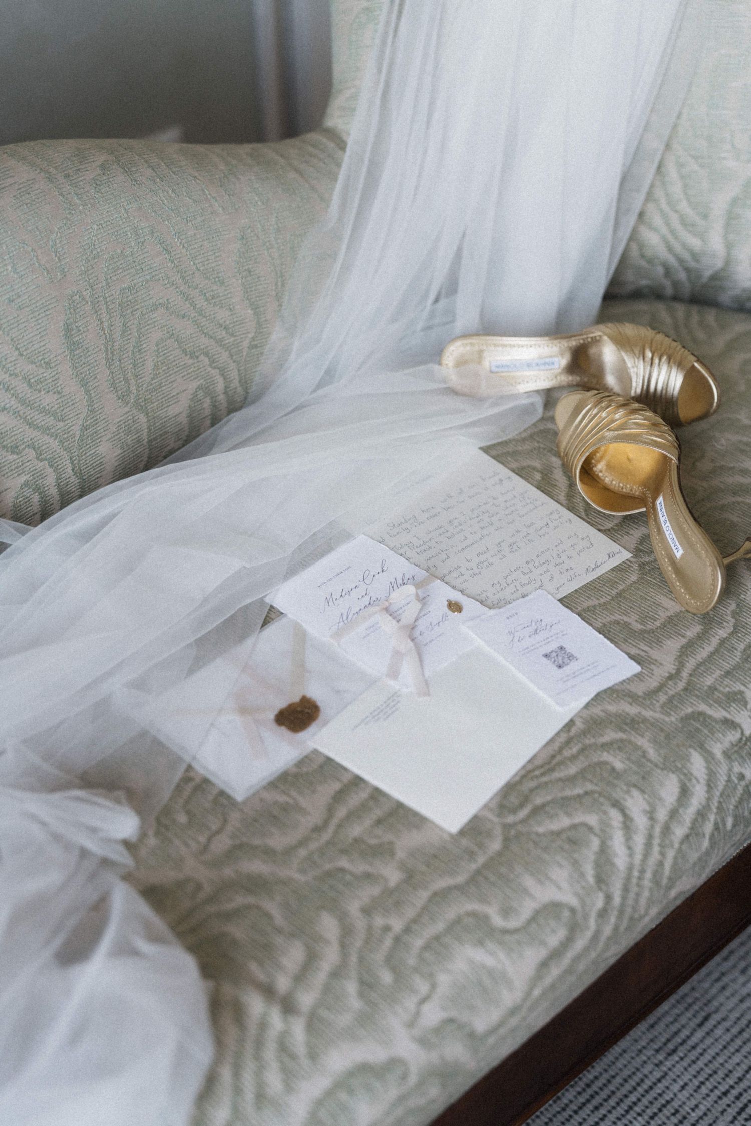 Bridal veil, gold heels, and wedding stationery with wax seal arranged on a quilted sofa.