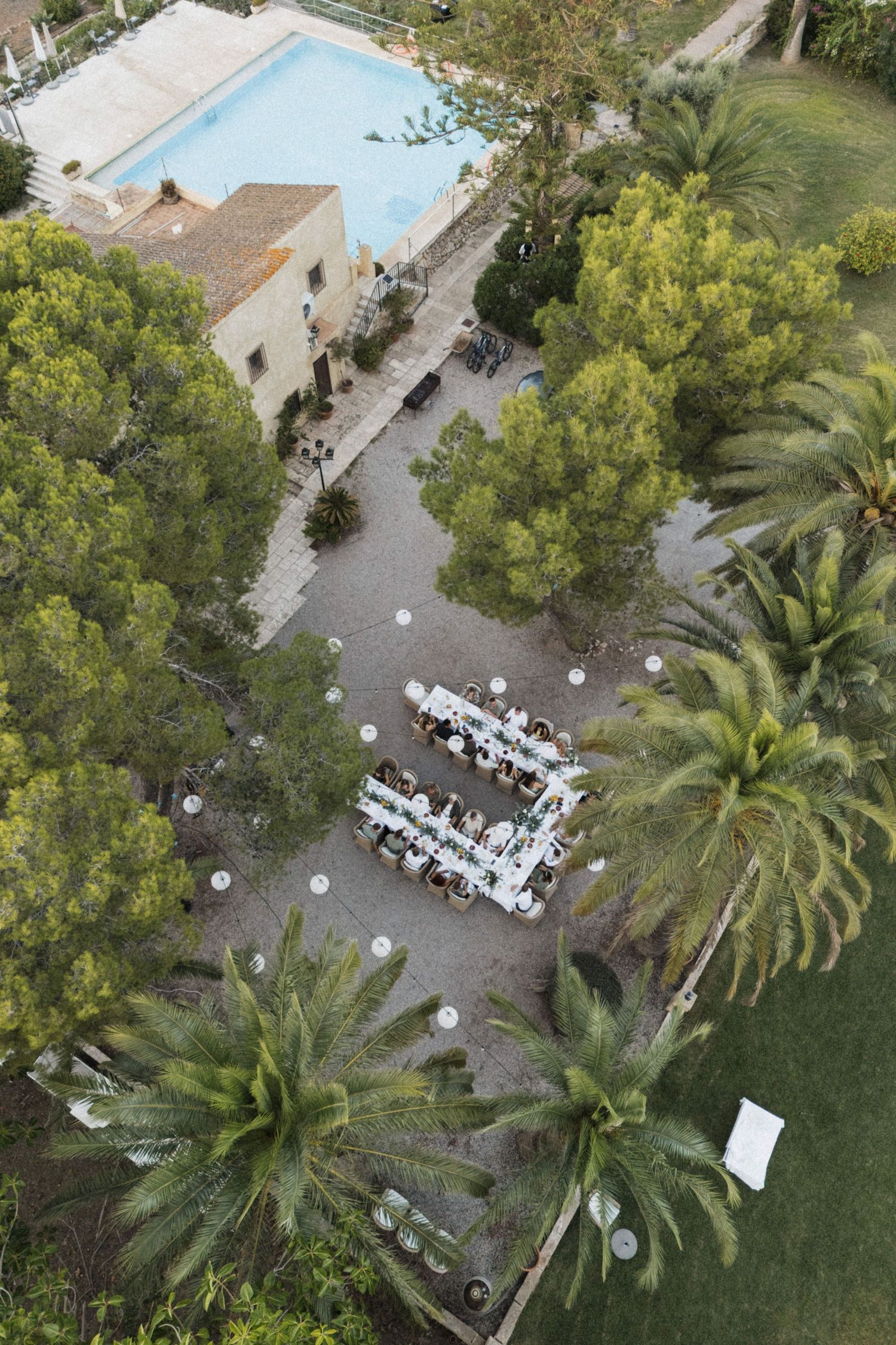 Aerial view of a wedding reception with long white tables set among palm trees near a pool.