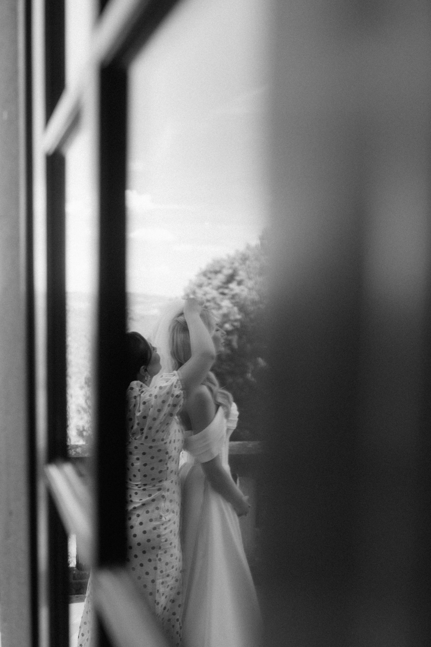 Black and white candid shot of bride and assistant reflected through a window outdoors.