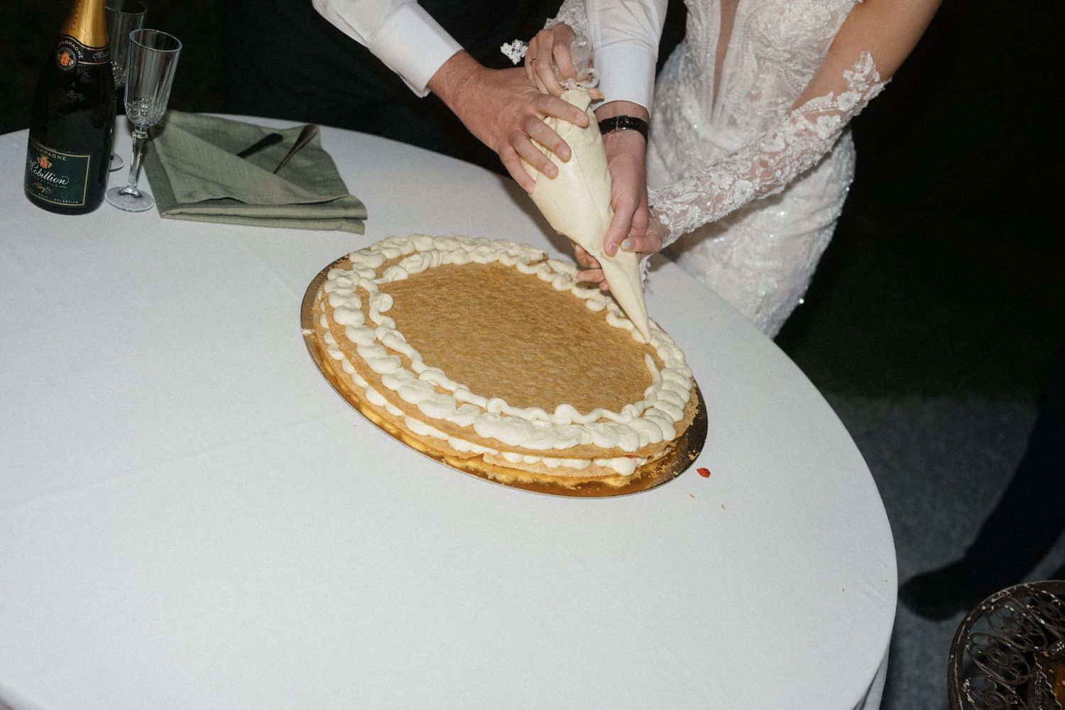 Bride decorating an oval wedding cake with cream piping at a white table during wedding reception.