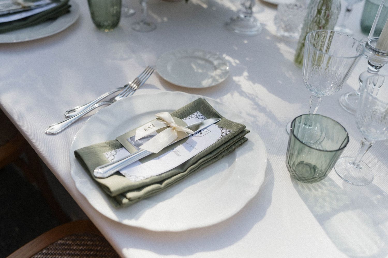 Elegant wedding place setting with sage green linen napkin, silverware, and glassware on a white tablecloth.
