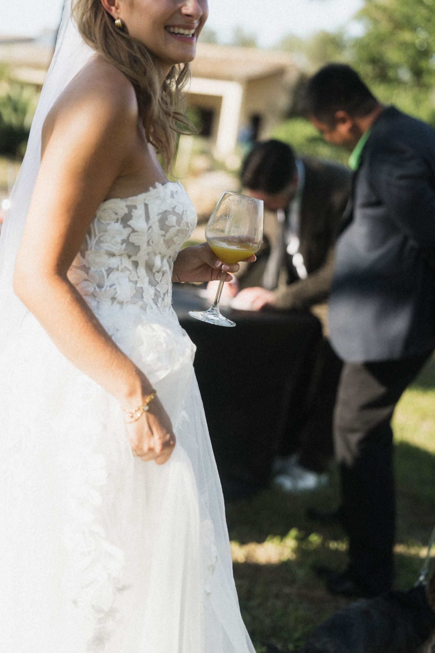 Elegant bride in strapless lace wedding gown holding a glass of white wine at outdoor reception.