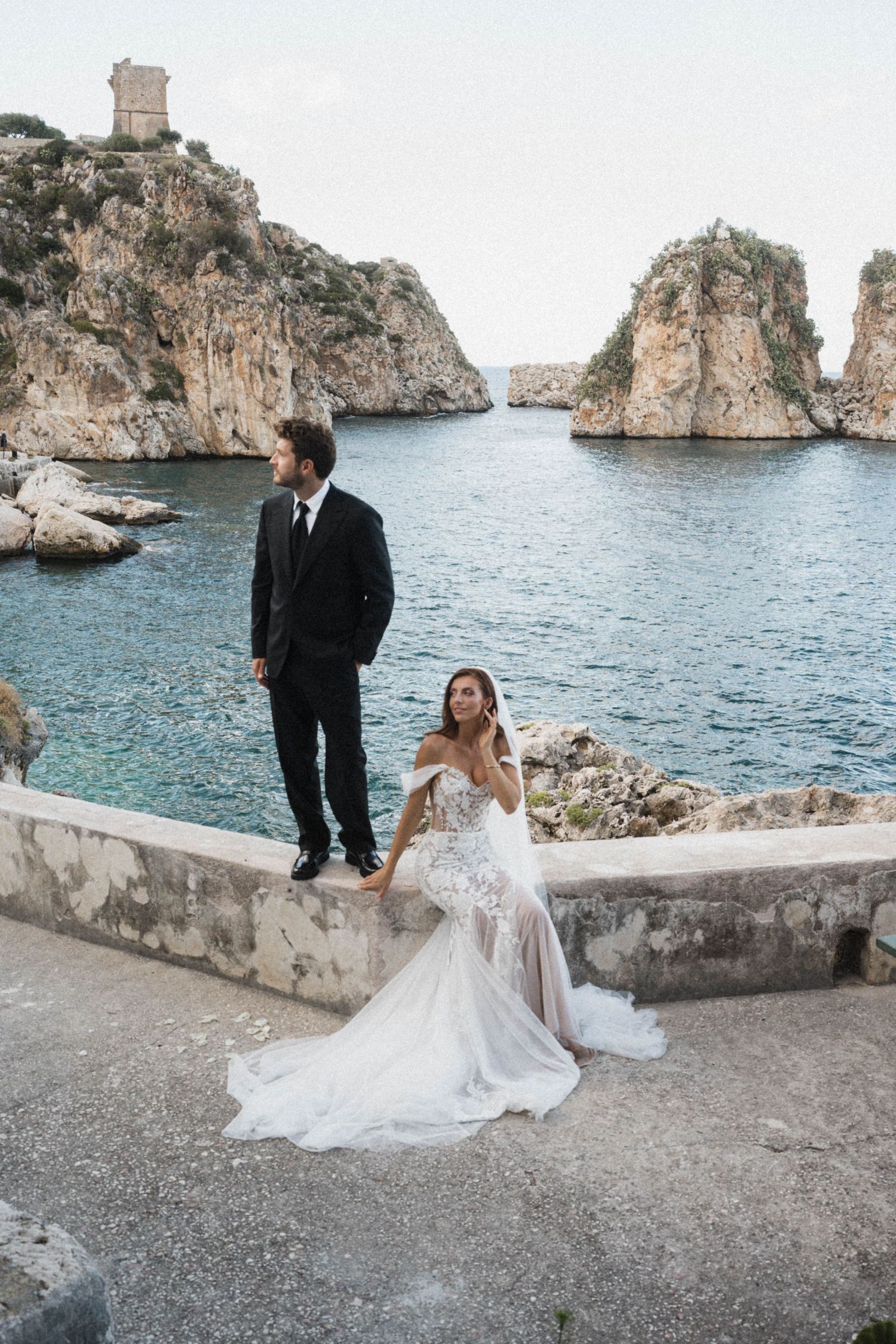 Bride in floral lace mermaid gown and groom in black suit posing by turquoise sea with rocky cliffs.