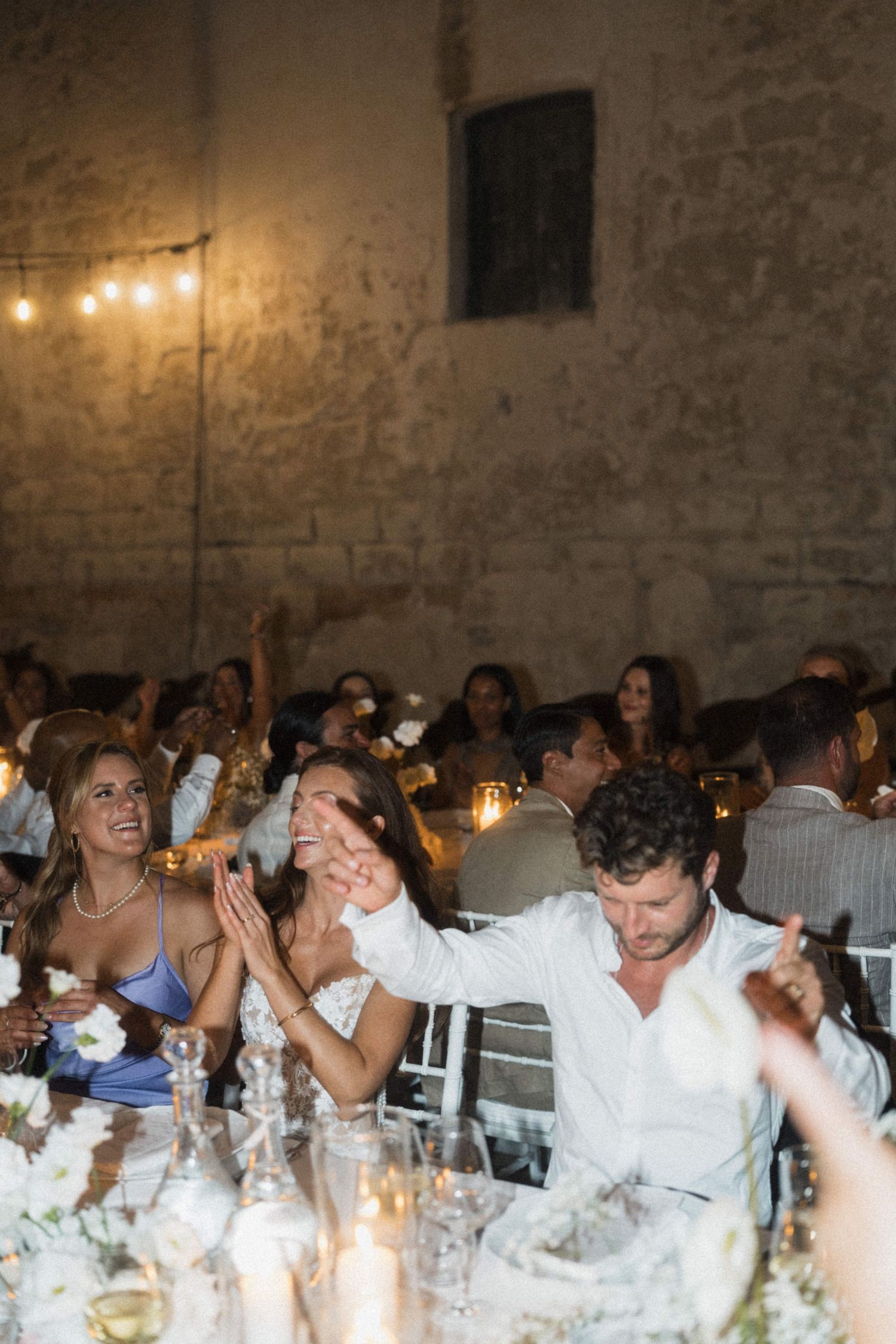 Guests clapping and celebrating at a candlelit wedding reception in a rustic stone venue with string lights.