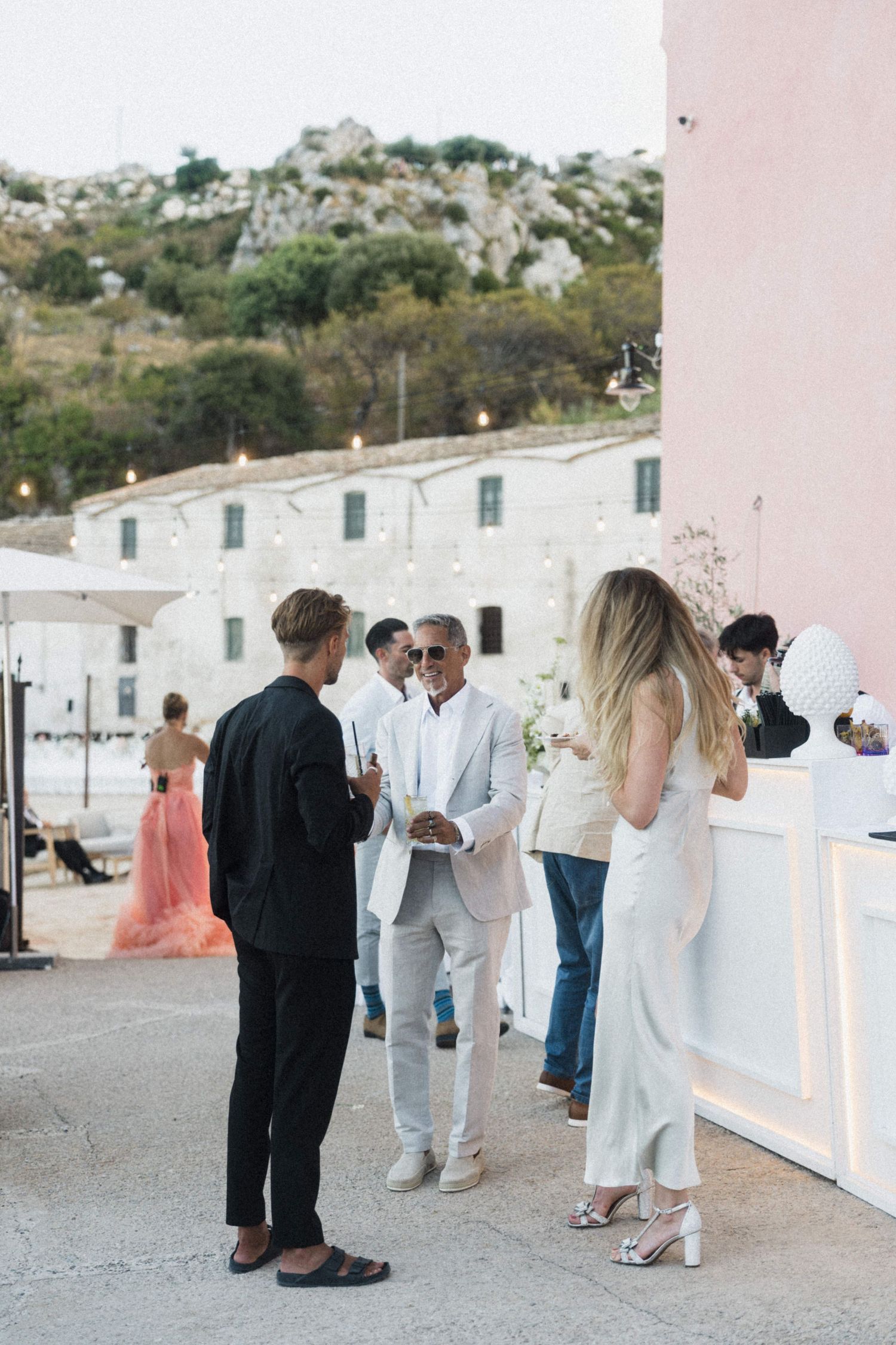 Wedding guests socializing on terrace near pink building with string lights and rocky hillside in background.
