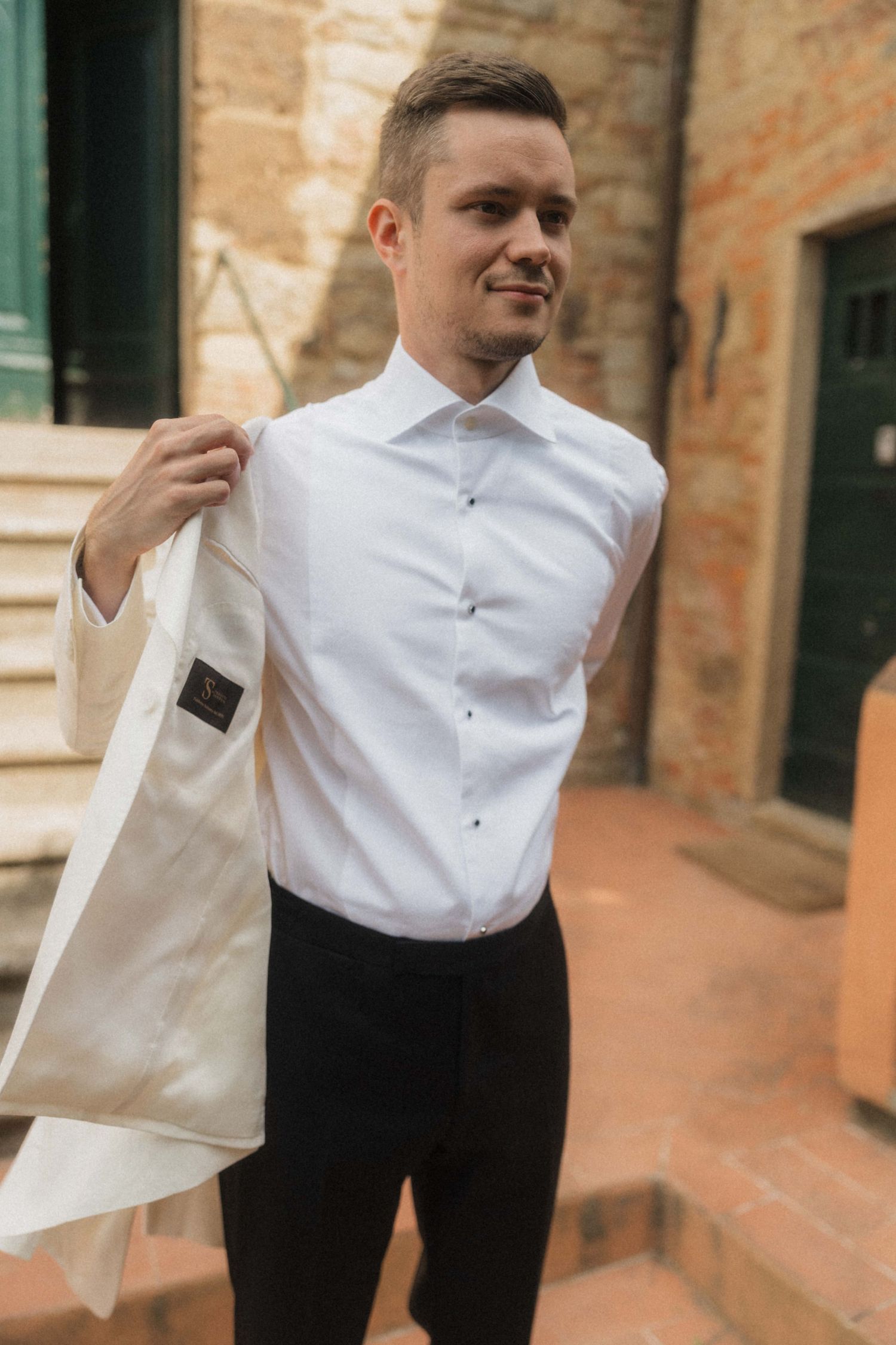 Groom in white dress shirt and black trousers putting on ivory wedding jacket outdoors at rustic venue.