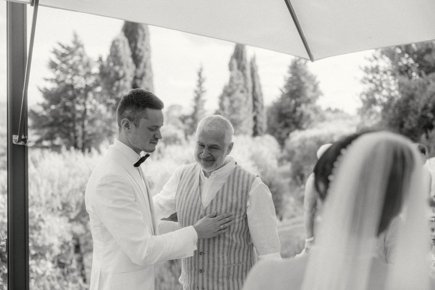 Black and white wedding ceremony photo showing groom shaking hands with officiant while bride stands nearby outdoors.