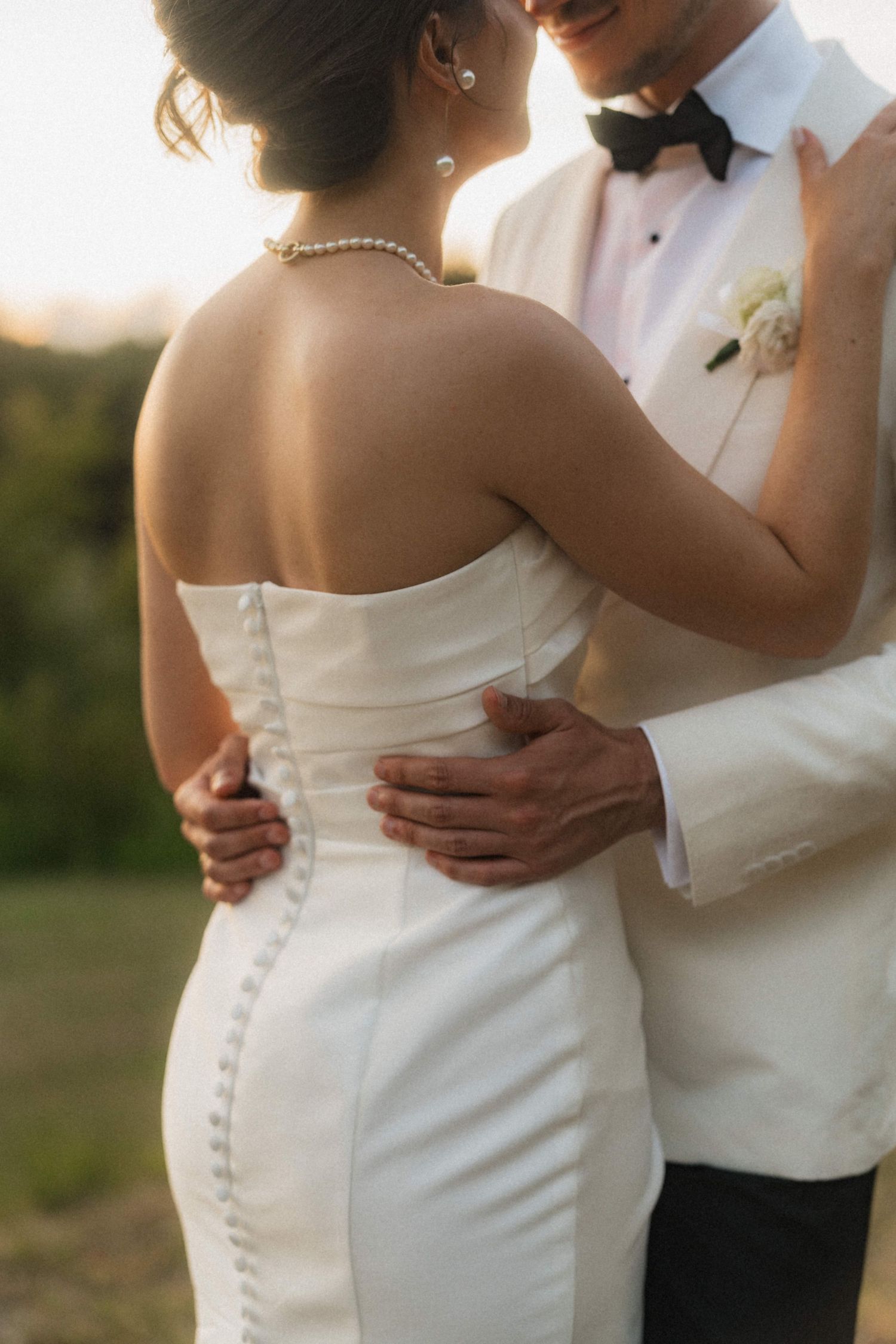 Bride in strapless button-back gown with pearl necklace embraced by smiling groom in white tuxedo at sunset.