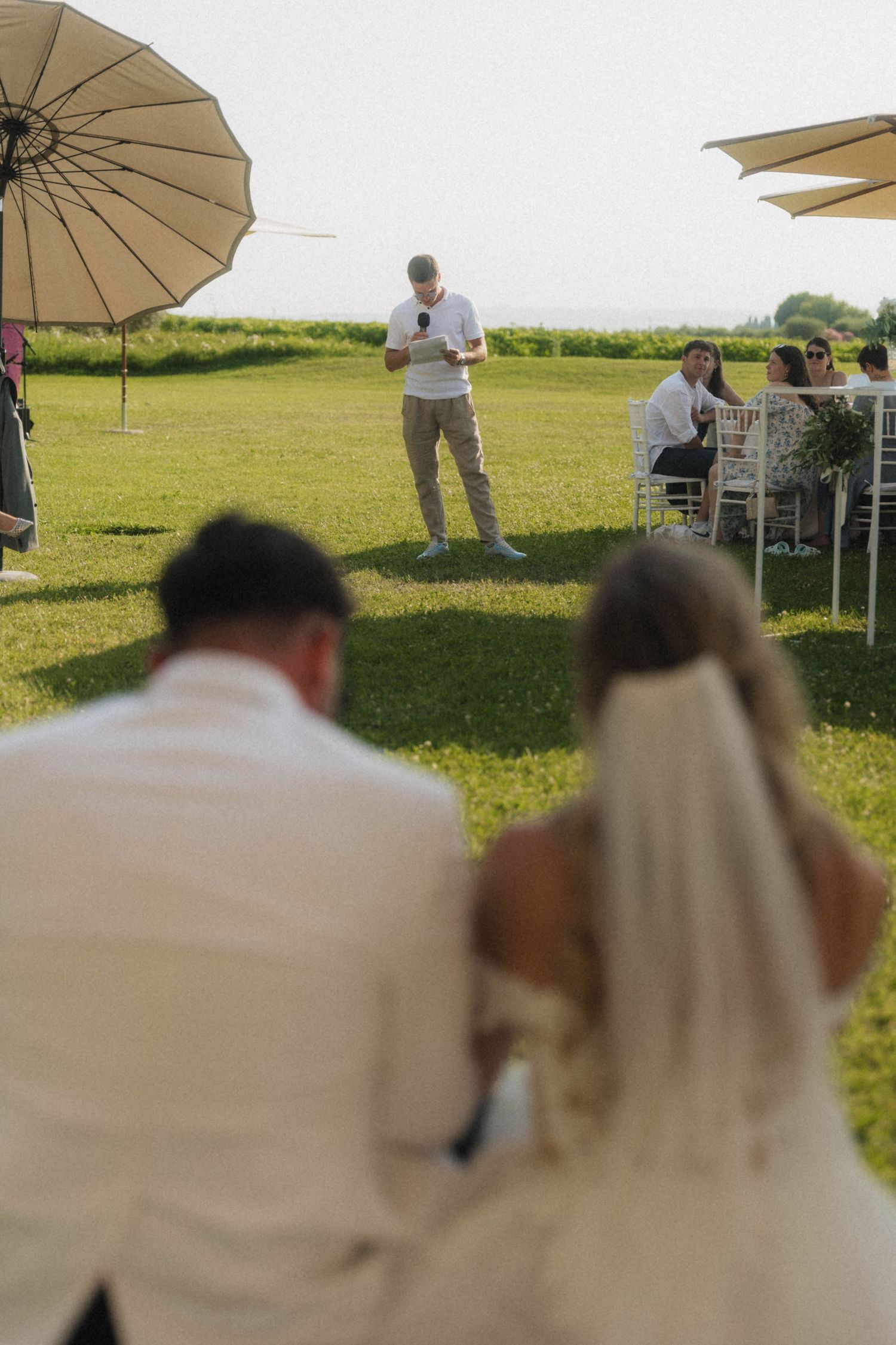 Bride and groom seated as guest delivers heartfelt speech at elegant outdoor wedding reception on green lawn.