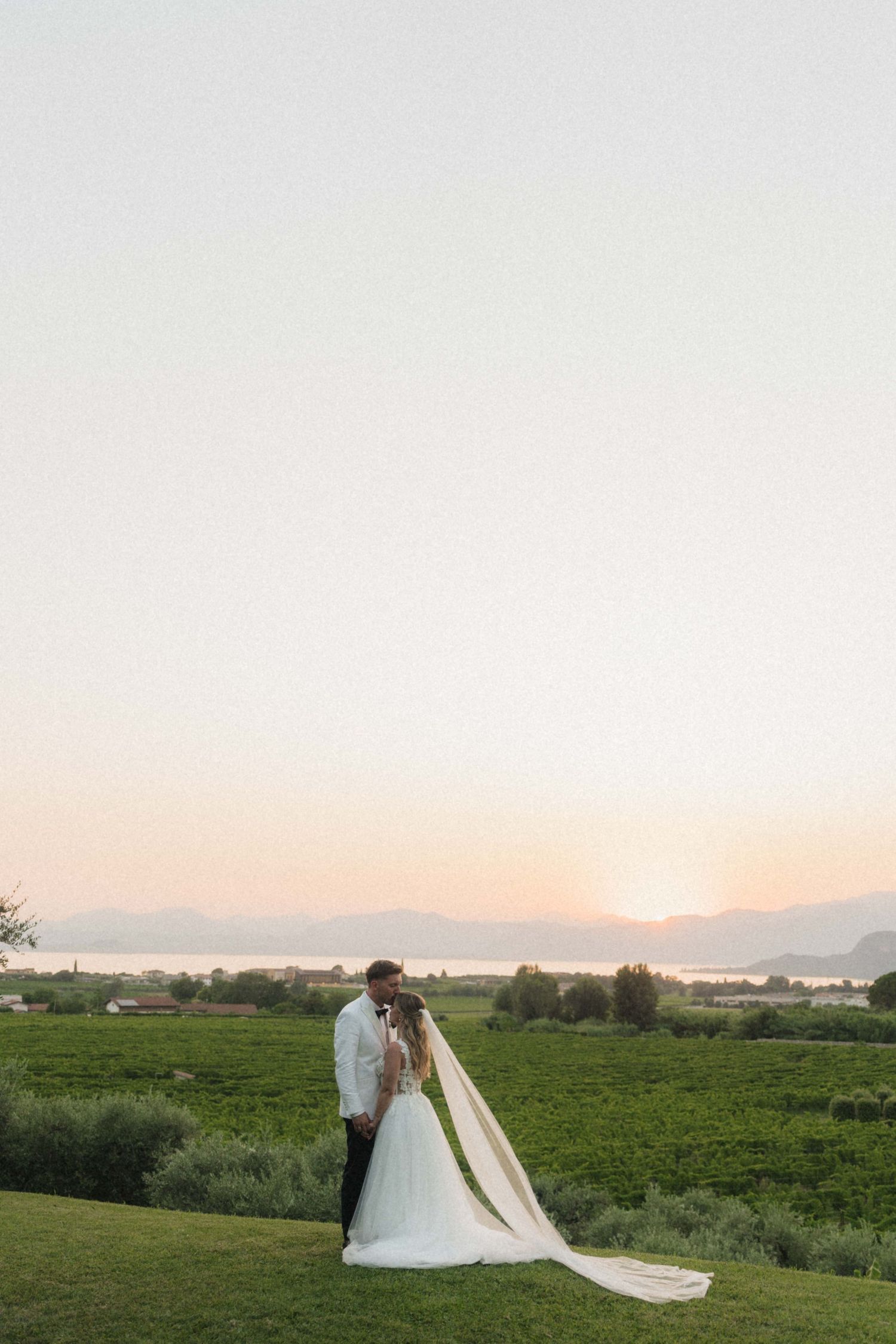 Bride and groom kissing at sunset overlooking scenic vineyard and mountain landscape.