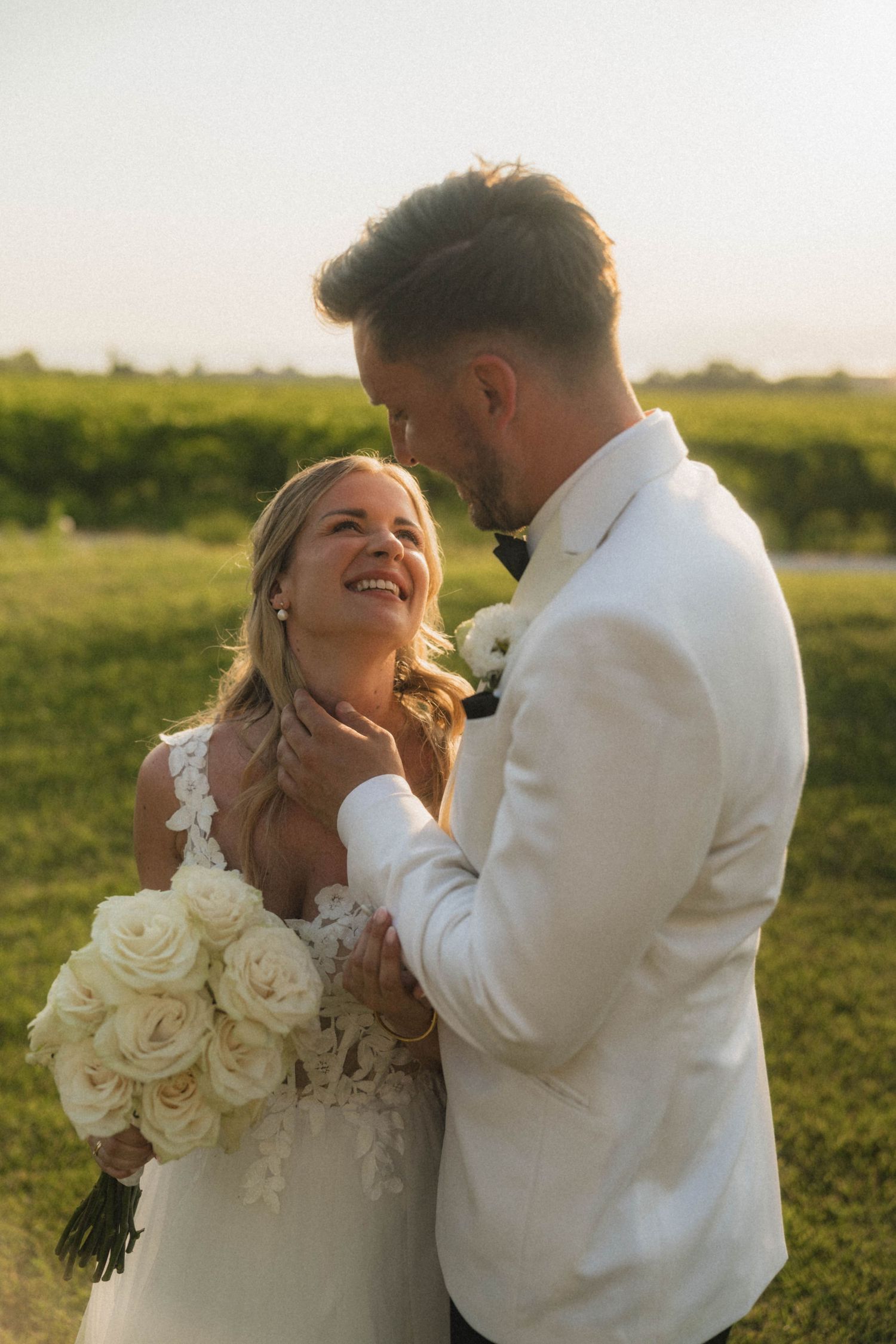 Smiling bride holding white rose bouquet gazes at groom in white jacket during golden hour.