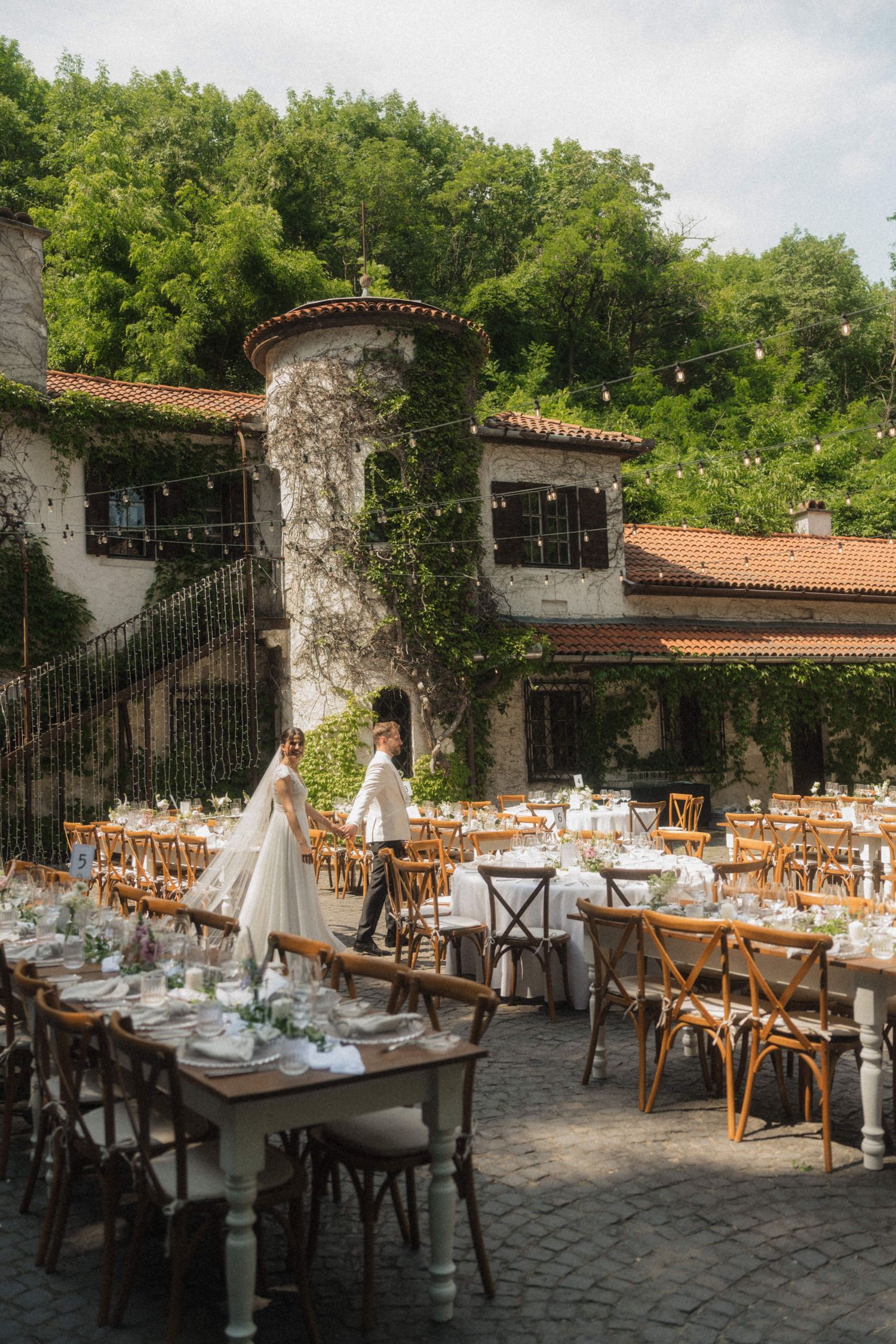 Outdoor wedding reception setup with wooden tables and chairs in a rustic villa courtyard with lush greenery.