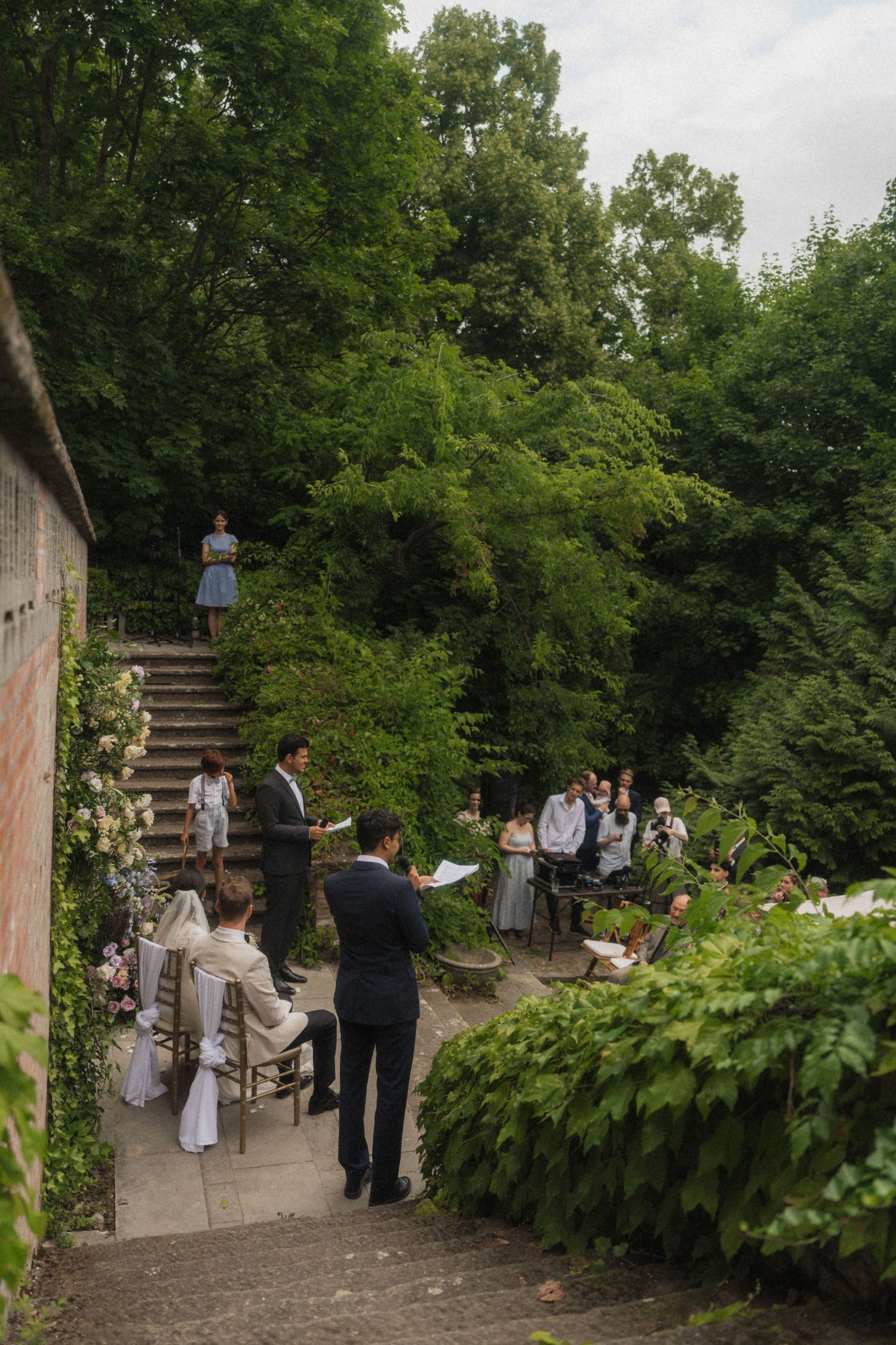 Outdoor wedding ceremony in a lush garden with guests seated among greenery and floral arrangements.