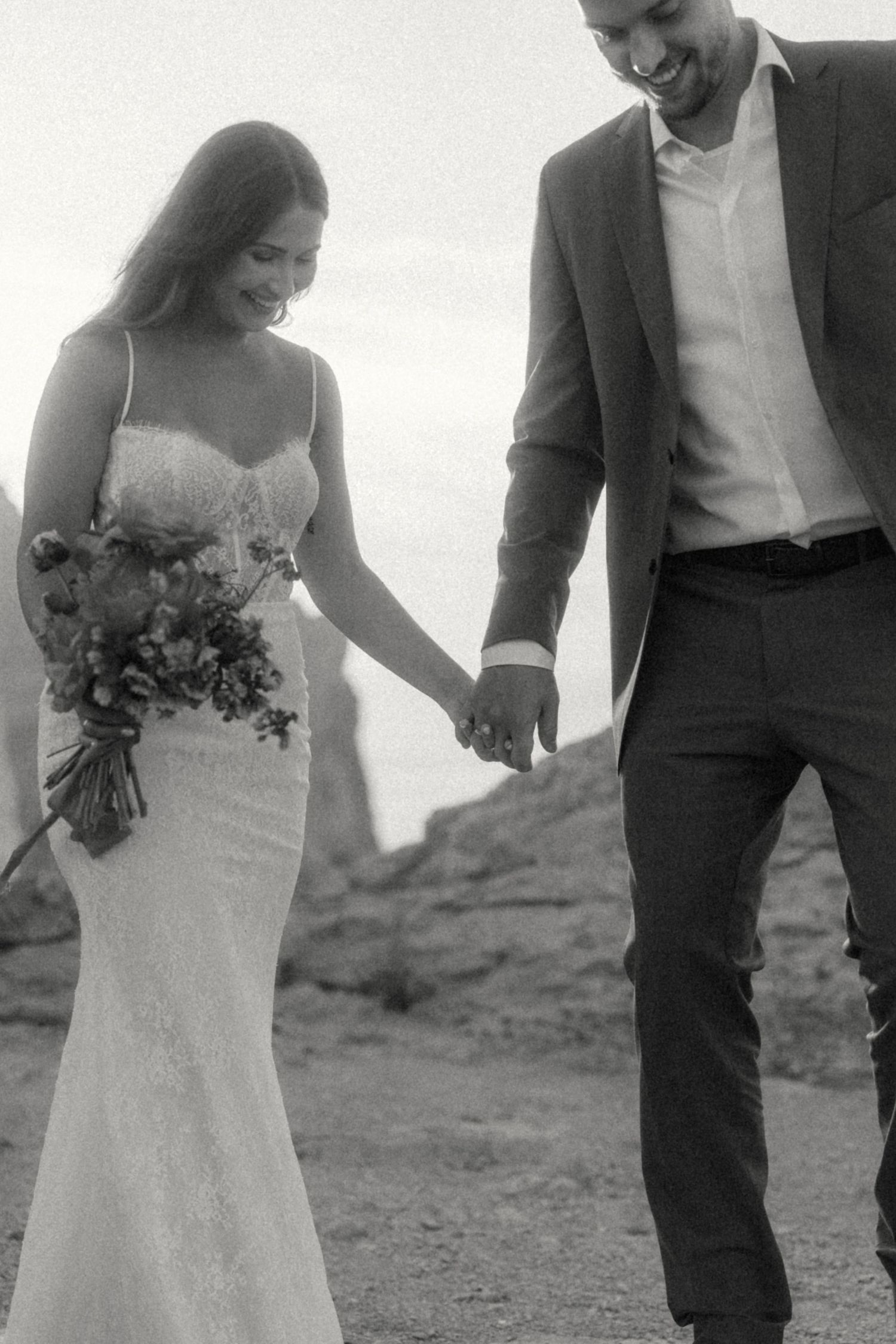 Black and white photo of smiling bride and groom holding hands walking outdoors, bride in spaghetti strap lace wedding gown.