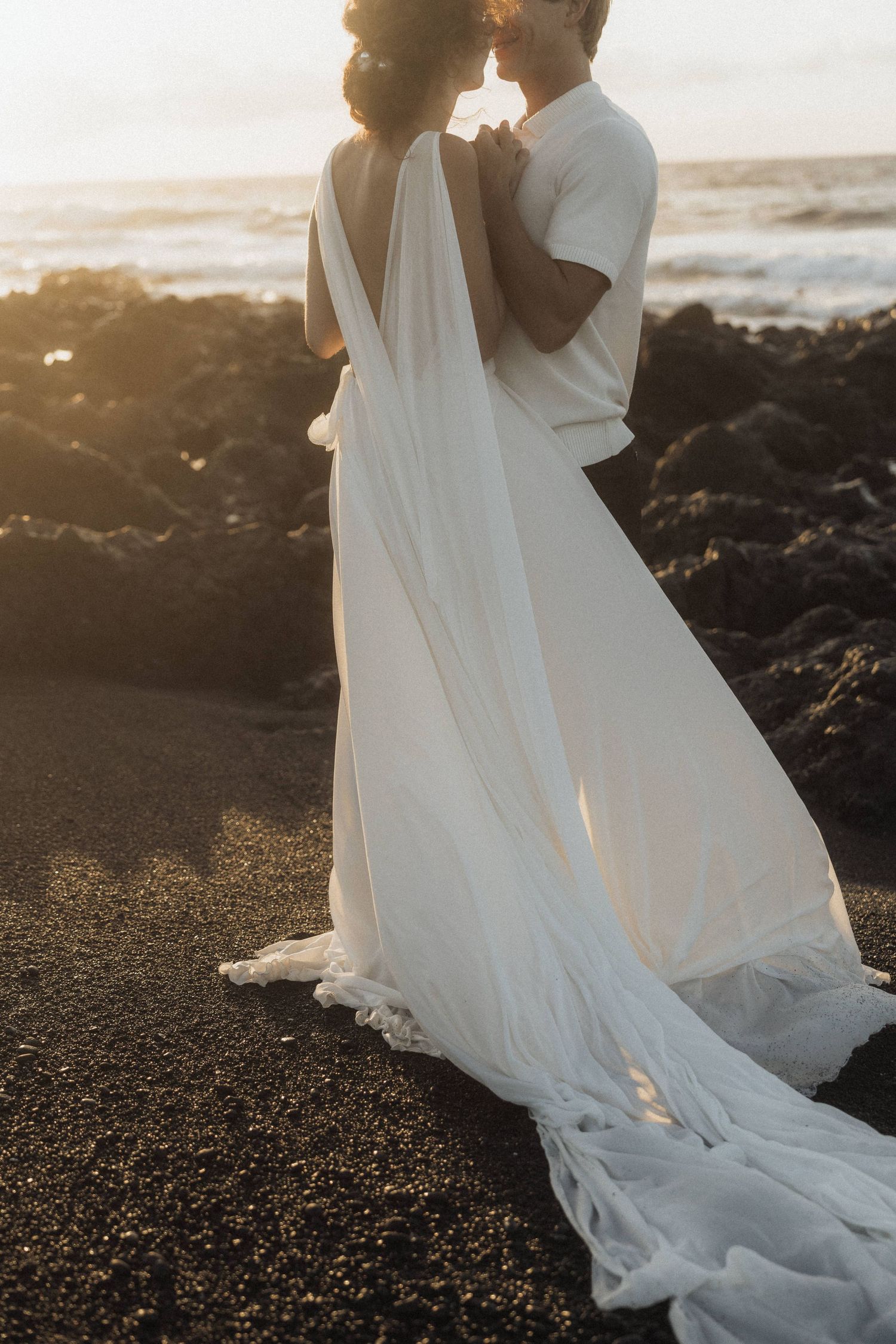 Bride in elegant backless white gown embracing groom on black sand beach at sunset.