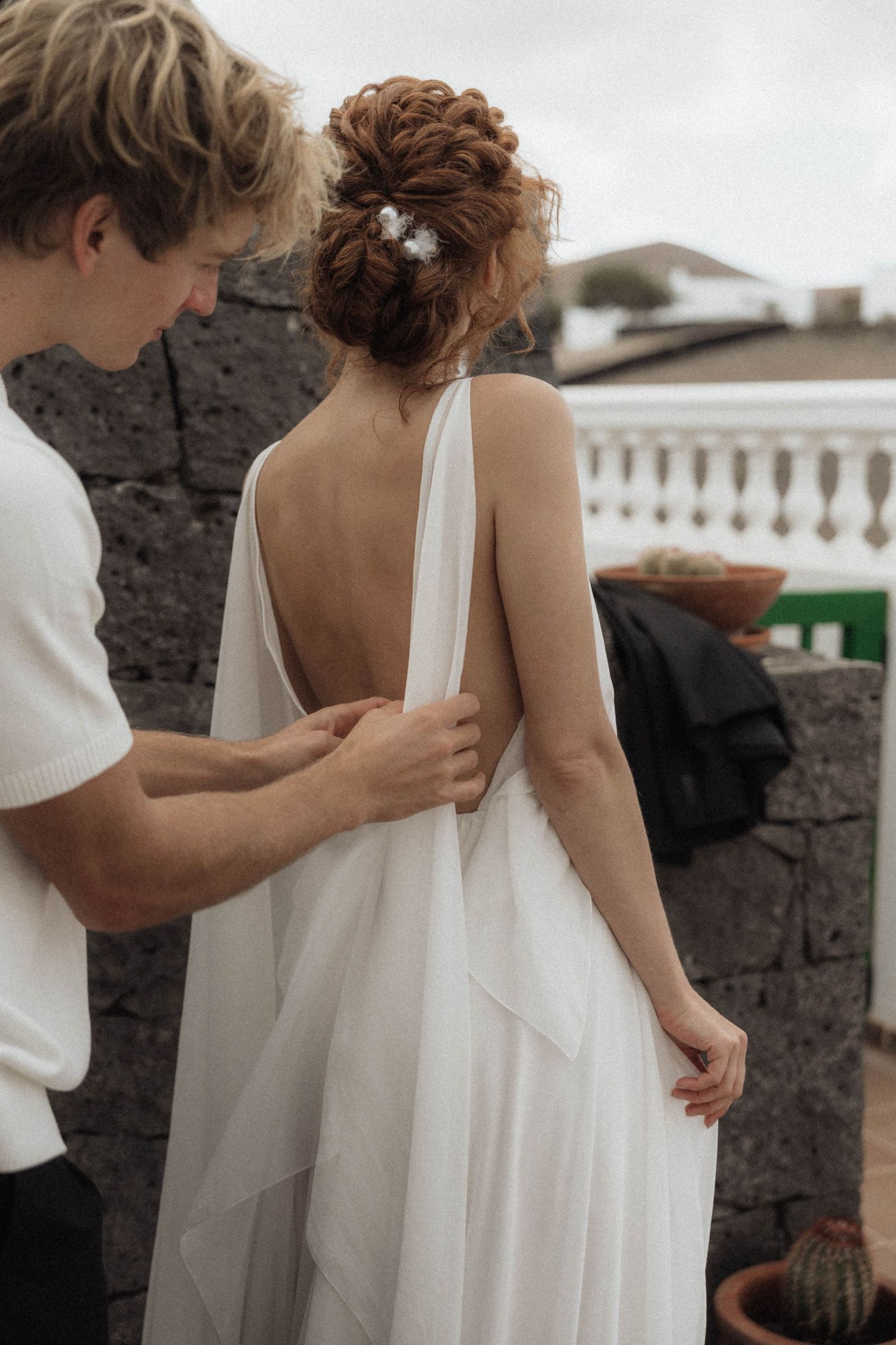 Groom adjusting bride's open-back white wedding dress outdoors near stone wall.