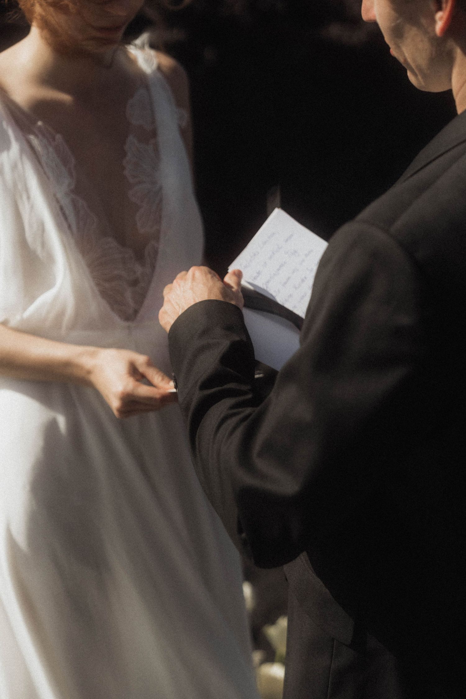 Groom reading wedding vows while holding bride's hand during outdoor ceremony, bride in white lace dress.