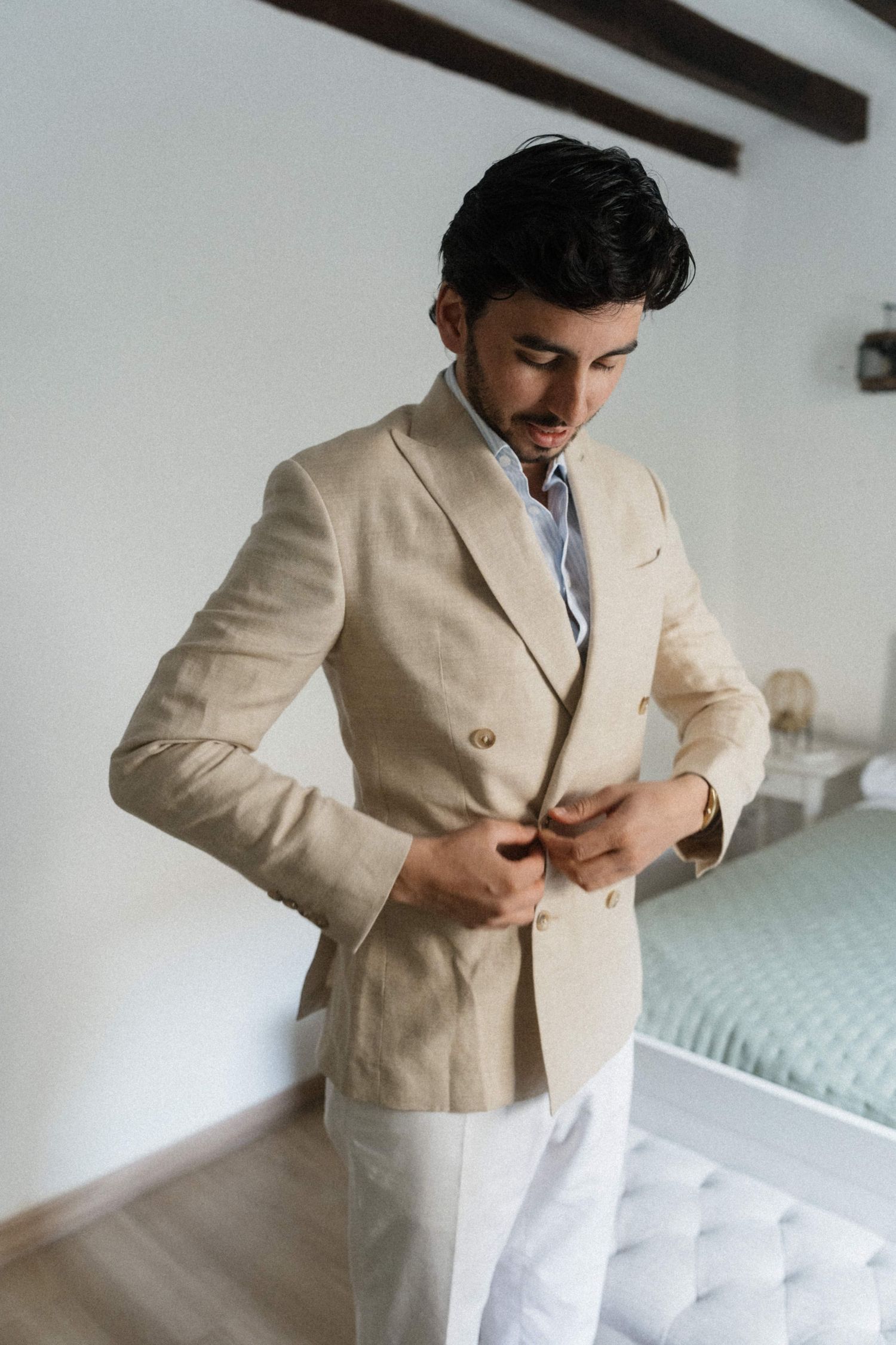 Groom buttoning a beige linen double-breasted blazer while getting dressed for his outdoor wedding ceremony.