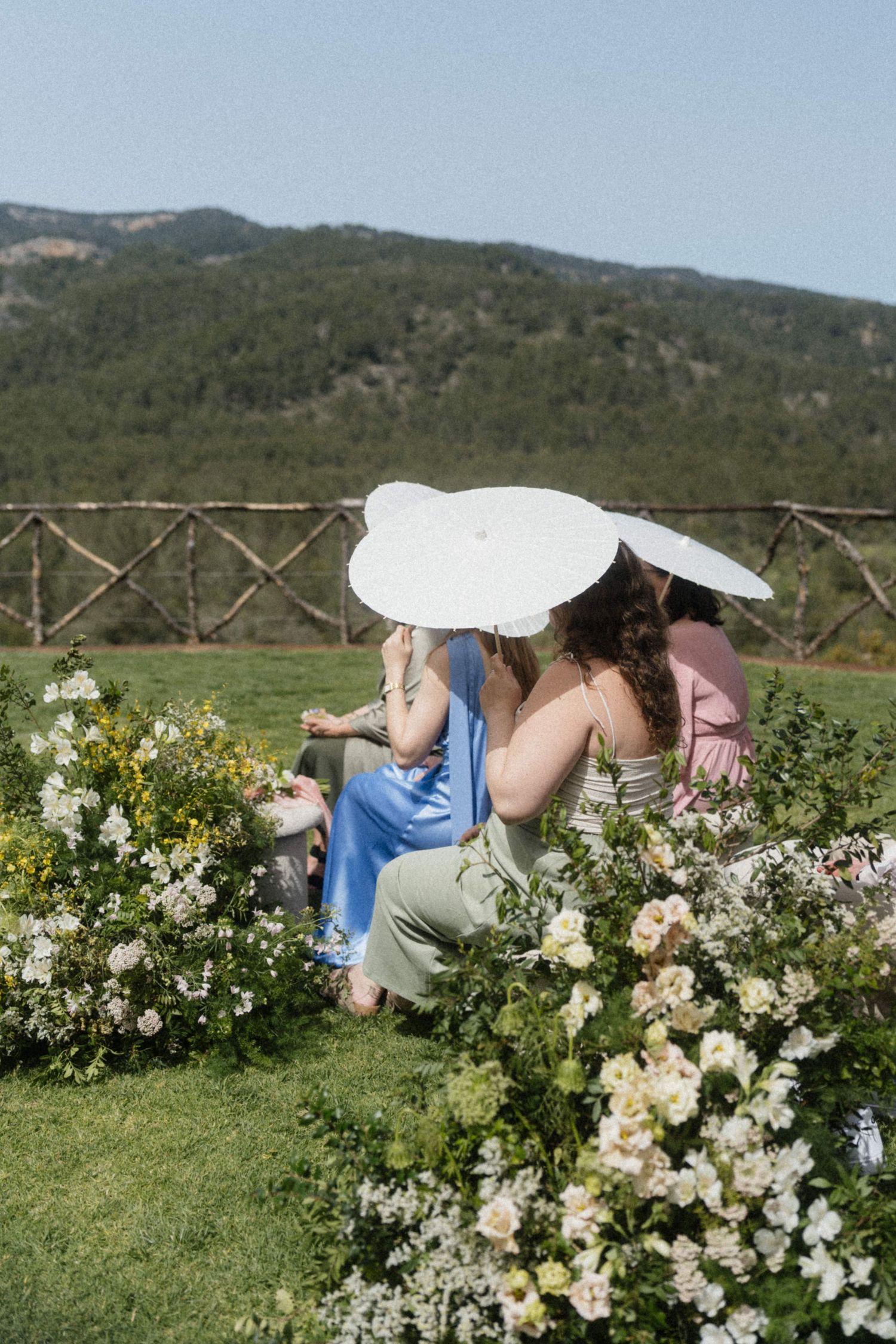Two women shielding from sun with white hats seated among white floral arrangements at outdoor wedding.