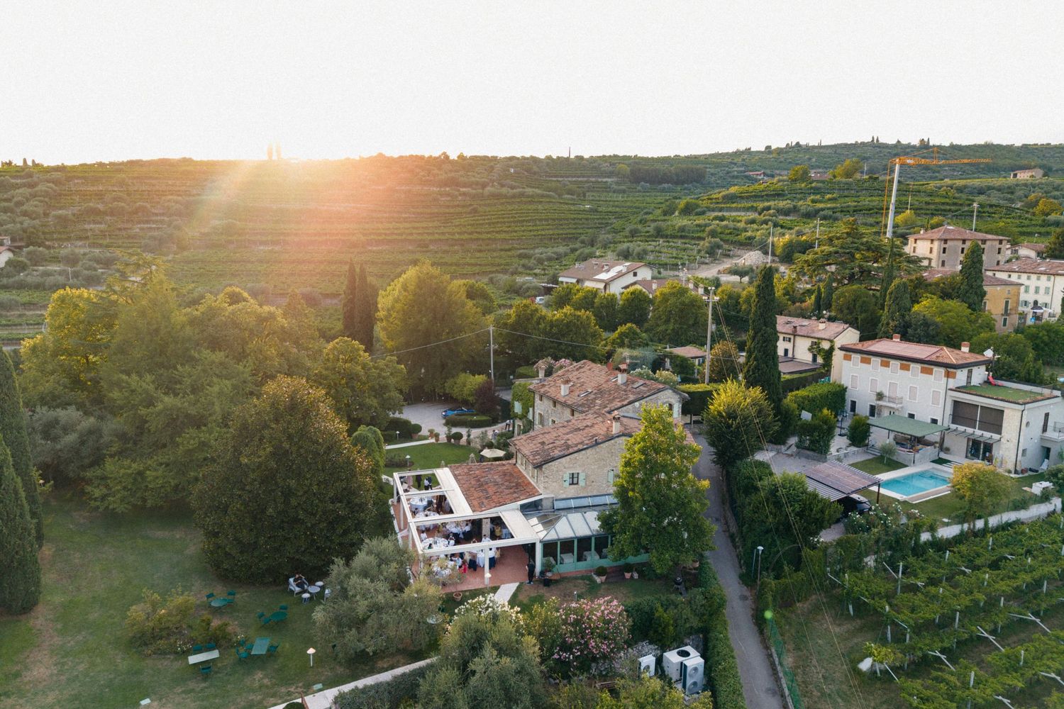 Aerial sunset view of a rustic Italian villa surrounded by lush vineyards and trees in the countryside.