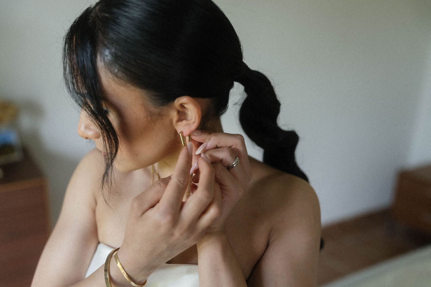 Bride putting on gold earring with ponytail hairstyle and gold bracelet on wedding day.