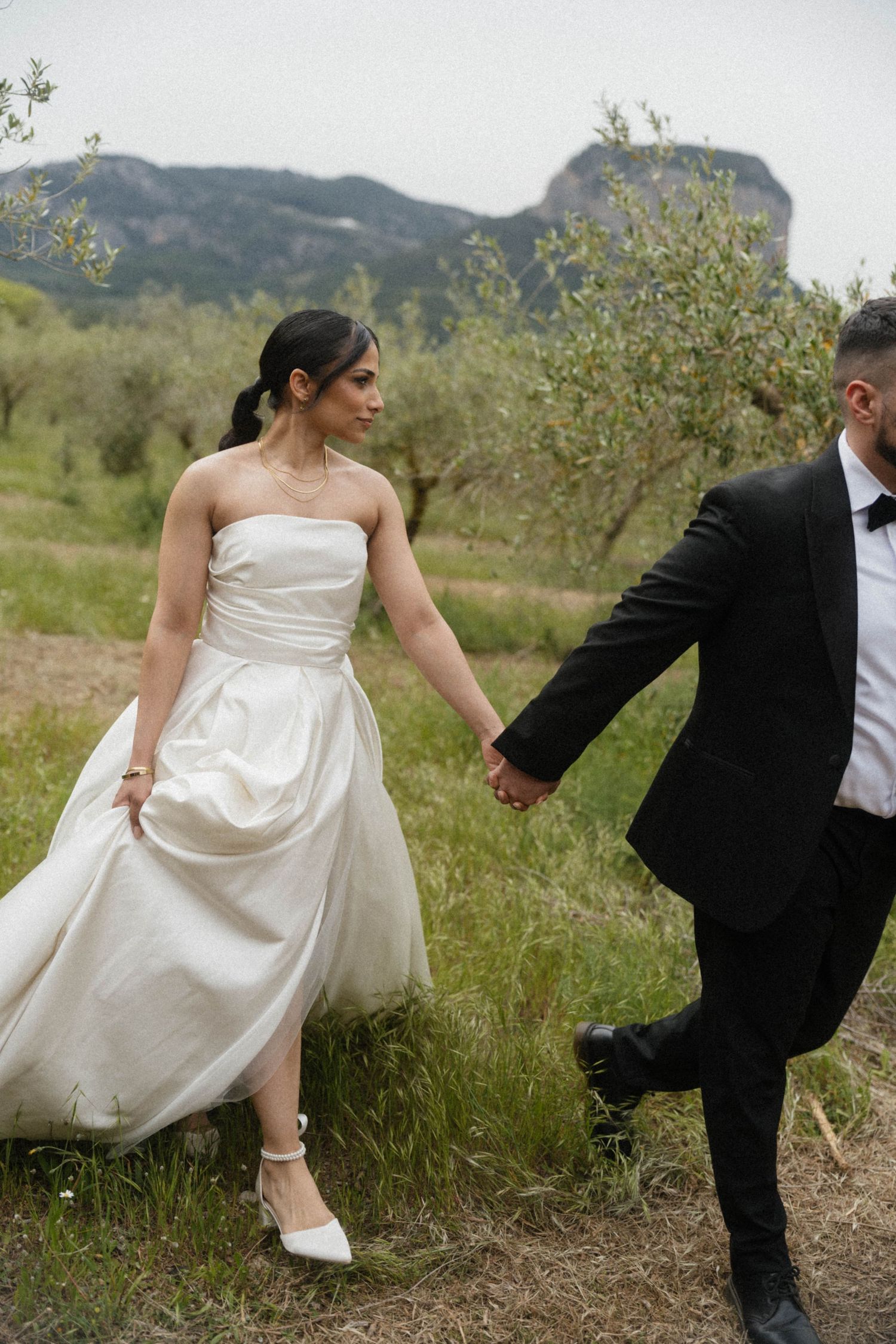 Bride in white strapless gown and groom in black tuxedo holding hands walking through olive grove.
