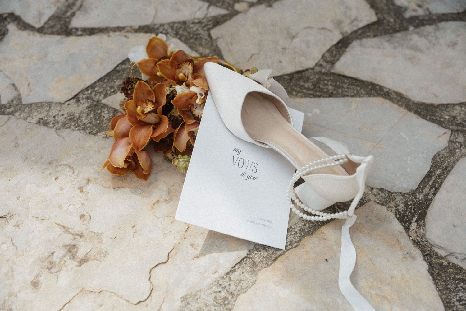 Wedding flat shoes beside vows booklet and dried floral arrangement on stone surface.