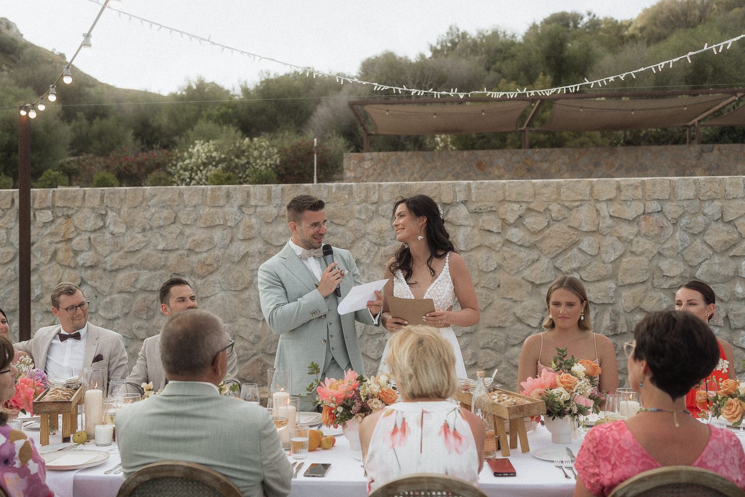 Bride and groom share vows at outdoor wedding reception with floral centerpieces and string lights.