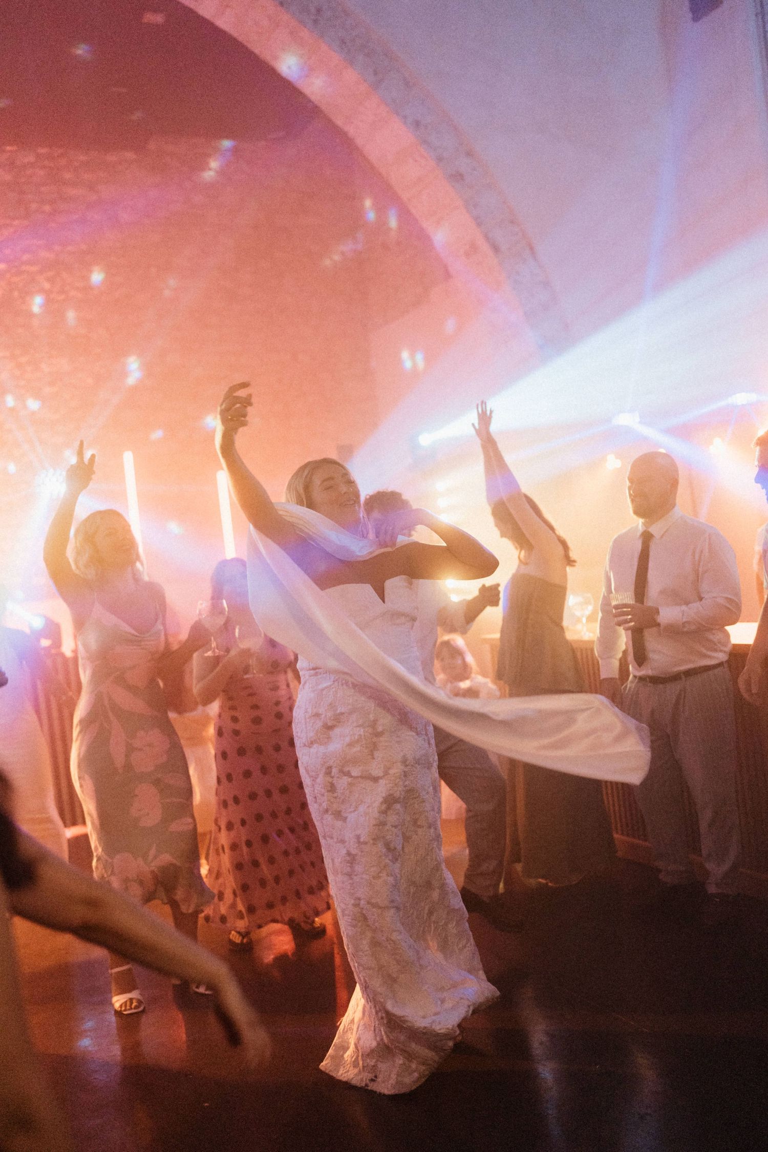 Bride in white lace gown dancing joyfully with guests at a wedding reception under colorful party lights.