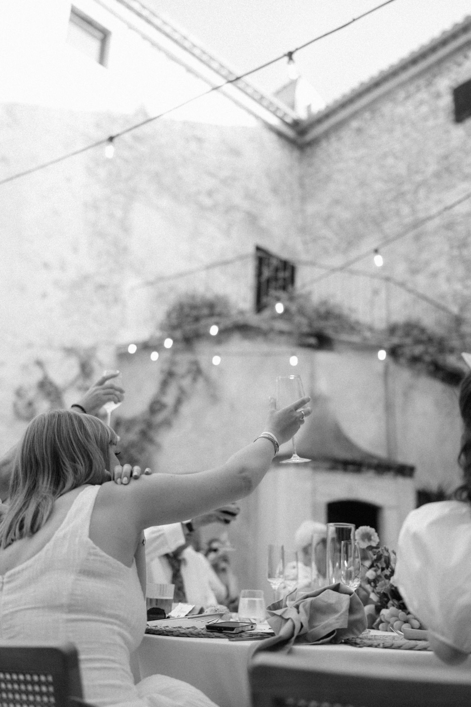 Black and white photo of wedding guests raising glasses in a toast at an outdoor reception dinner.
