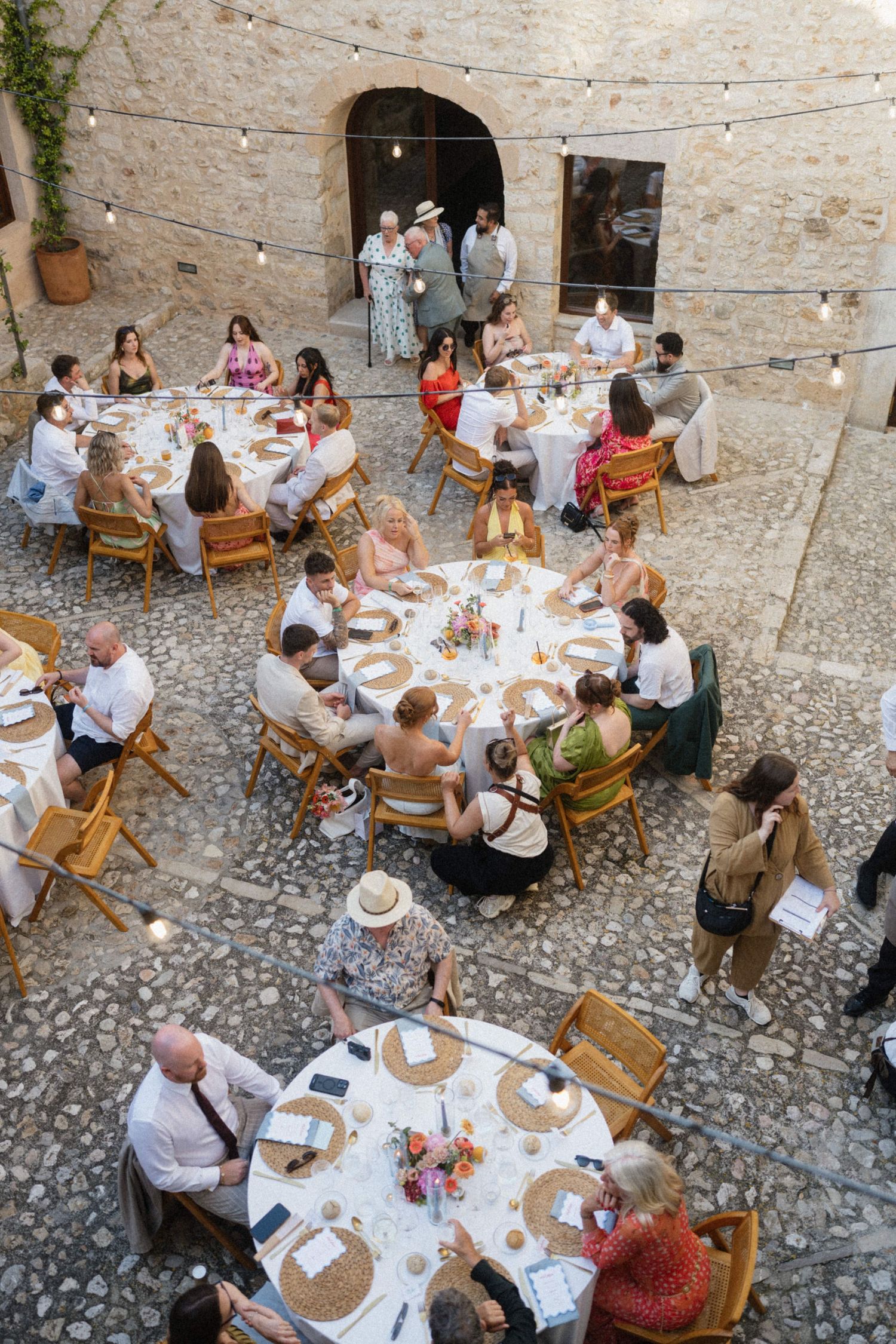 Aerial view of wedding guests dining at round tables in a rustic stone courtyard with string lights.