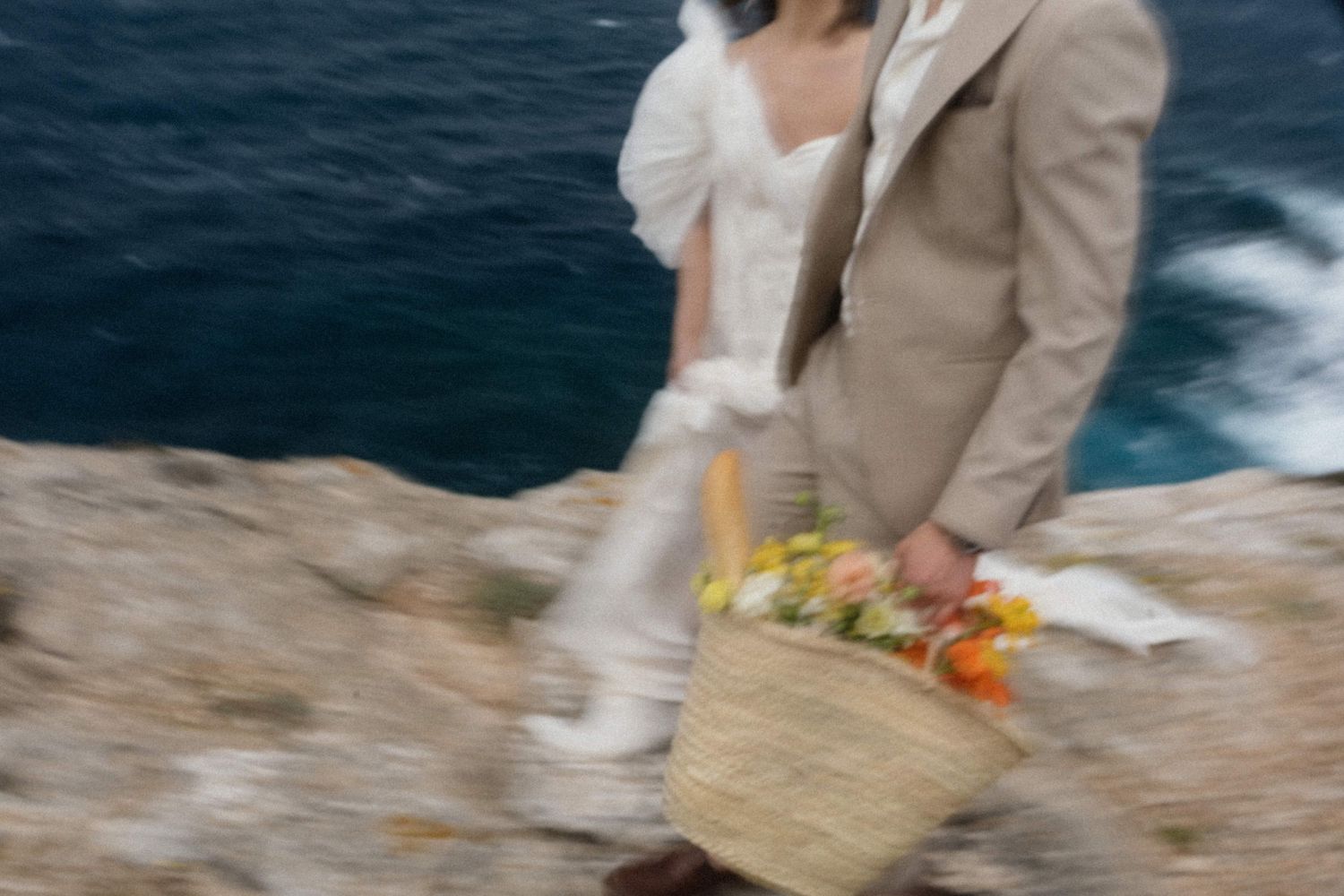 Blurred motion shot of a couple in wedding attire walking on rocky cliffs by the ocean, carrying a floral basket.