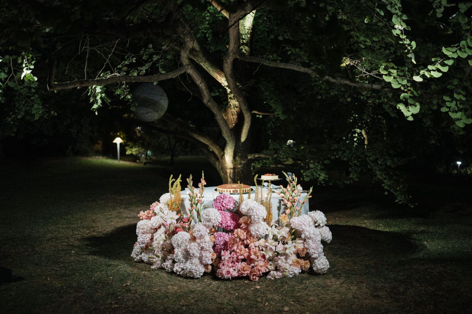 Romantic outdoor wedding sweetheart table adorned with lush pink floral arrangements under a tree at night.
