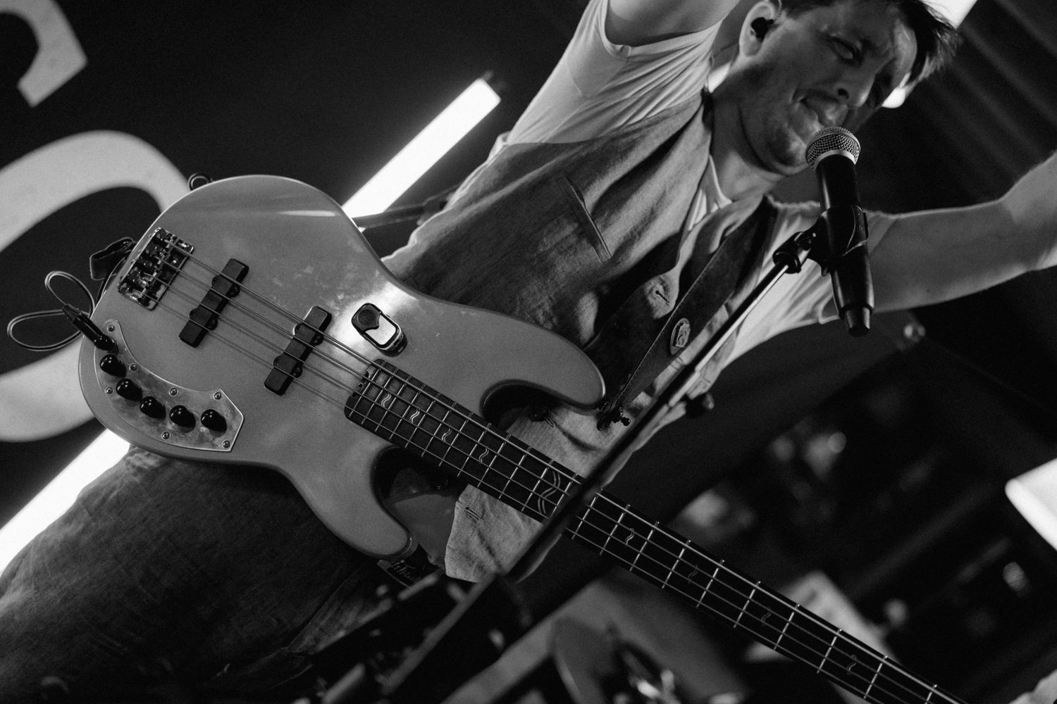 Black and white photo of a musician playing bass guitar on stage near a microphone.