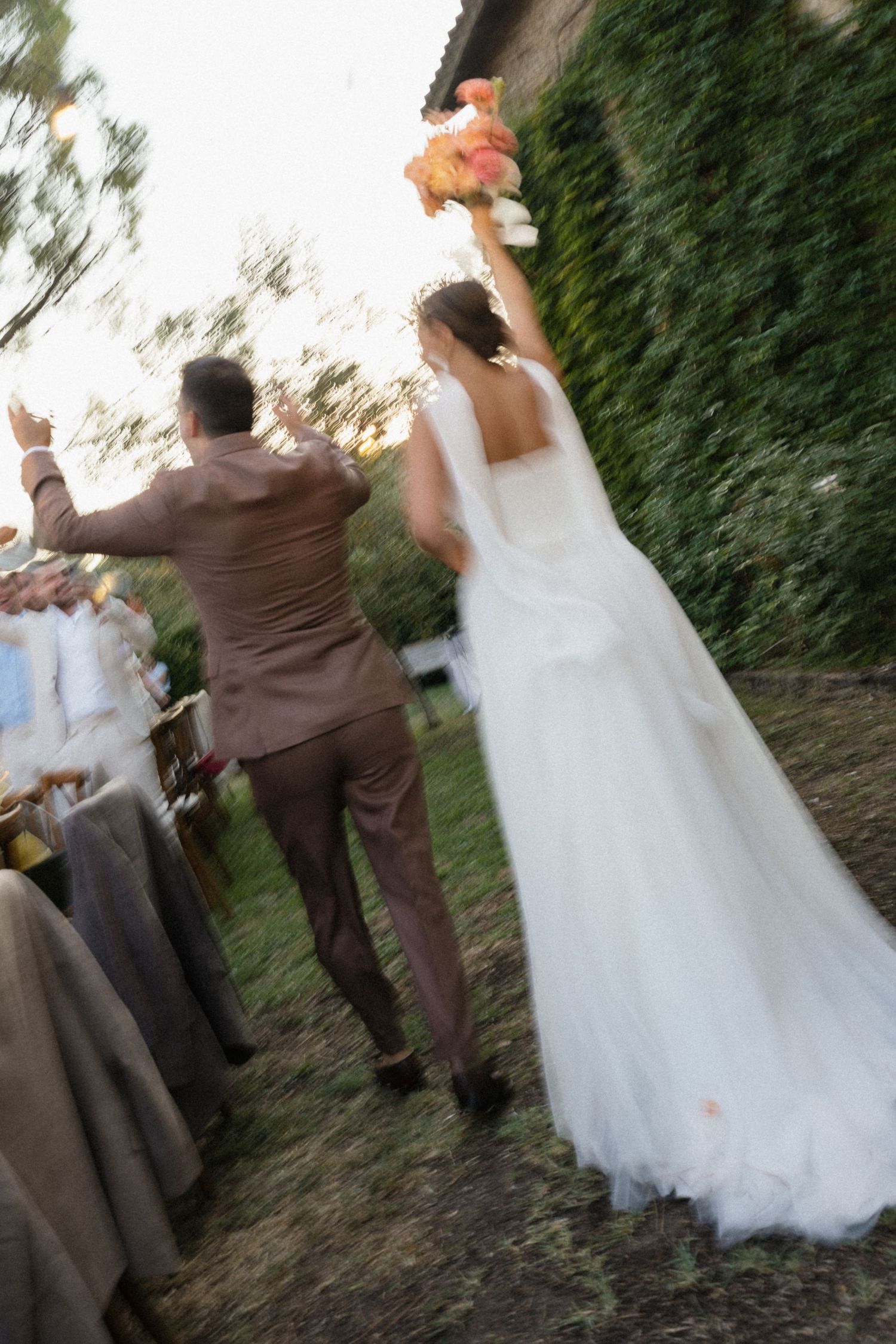 Bride and groom walking away, arms raised in celebration, bride in white gown holding bouquet, groom in tan suit.
