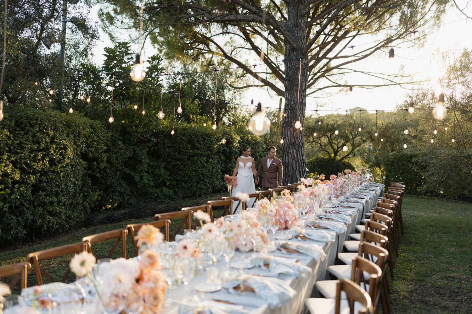 Elegant outdoor wedding reception with long dining table, floral centerpieces, hanging lights, and couple walking among lush greenery.