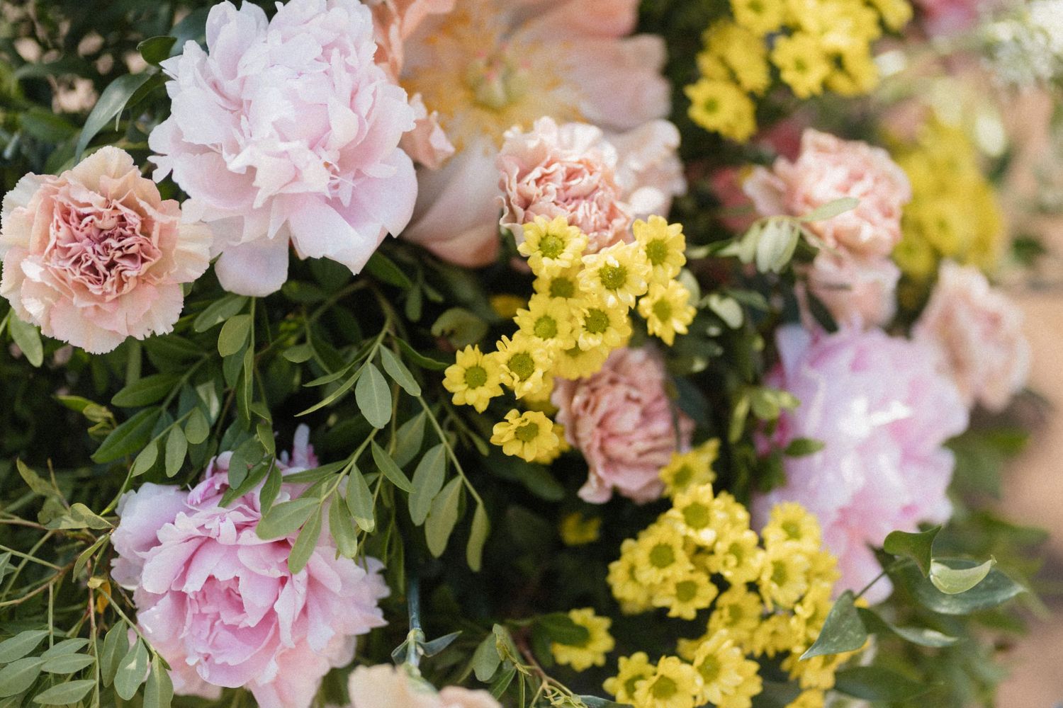 Lush floral arrangement featuring pink peonies, peach carnations, and yellow daisies with green foliage.