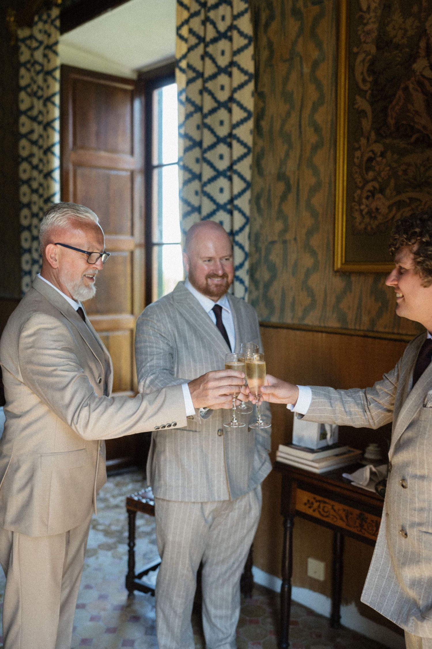 Three men in suits toasting with champagne glasses in an elegant room with patterned wallpaper.